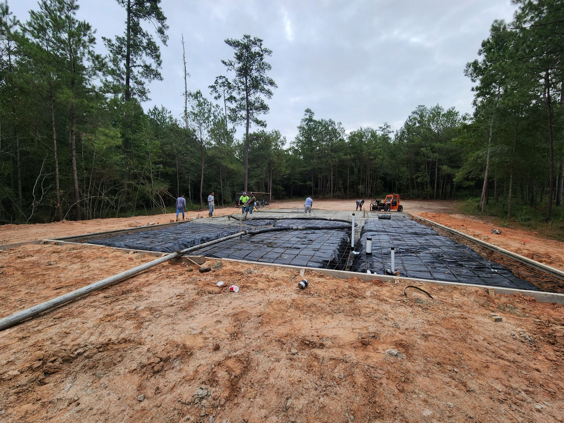 Construction site with workers, black material, and pipes in a cleared area surrounded by trees.