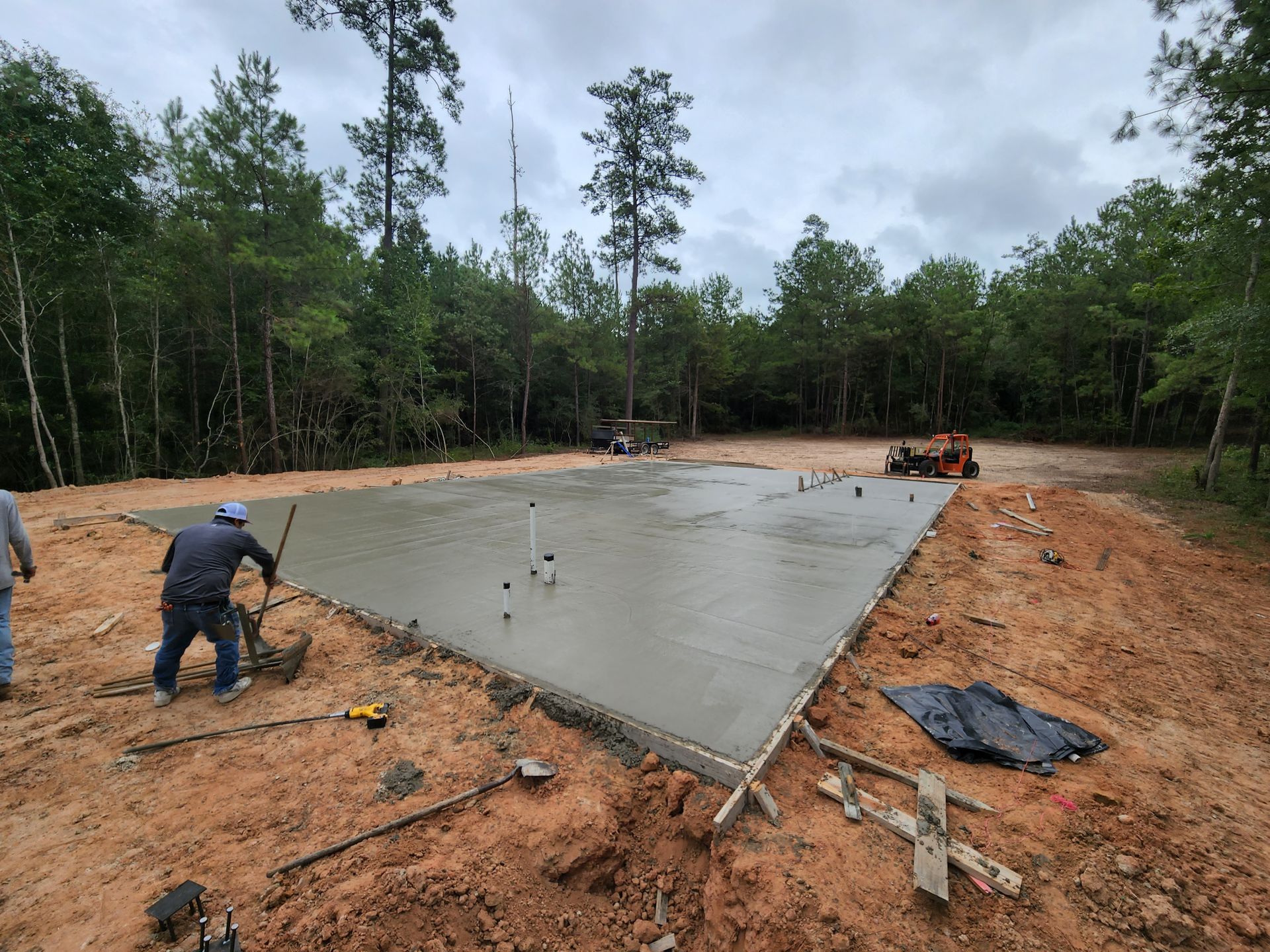 Workers pouring concrete foundation in a clearing surrounded by trees under a cloudy sky.