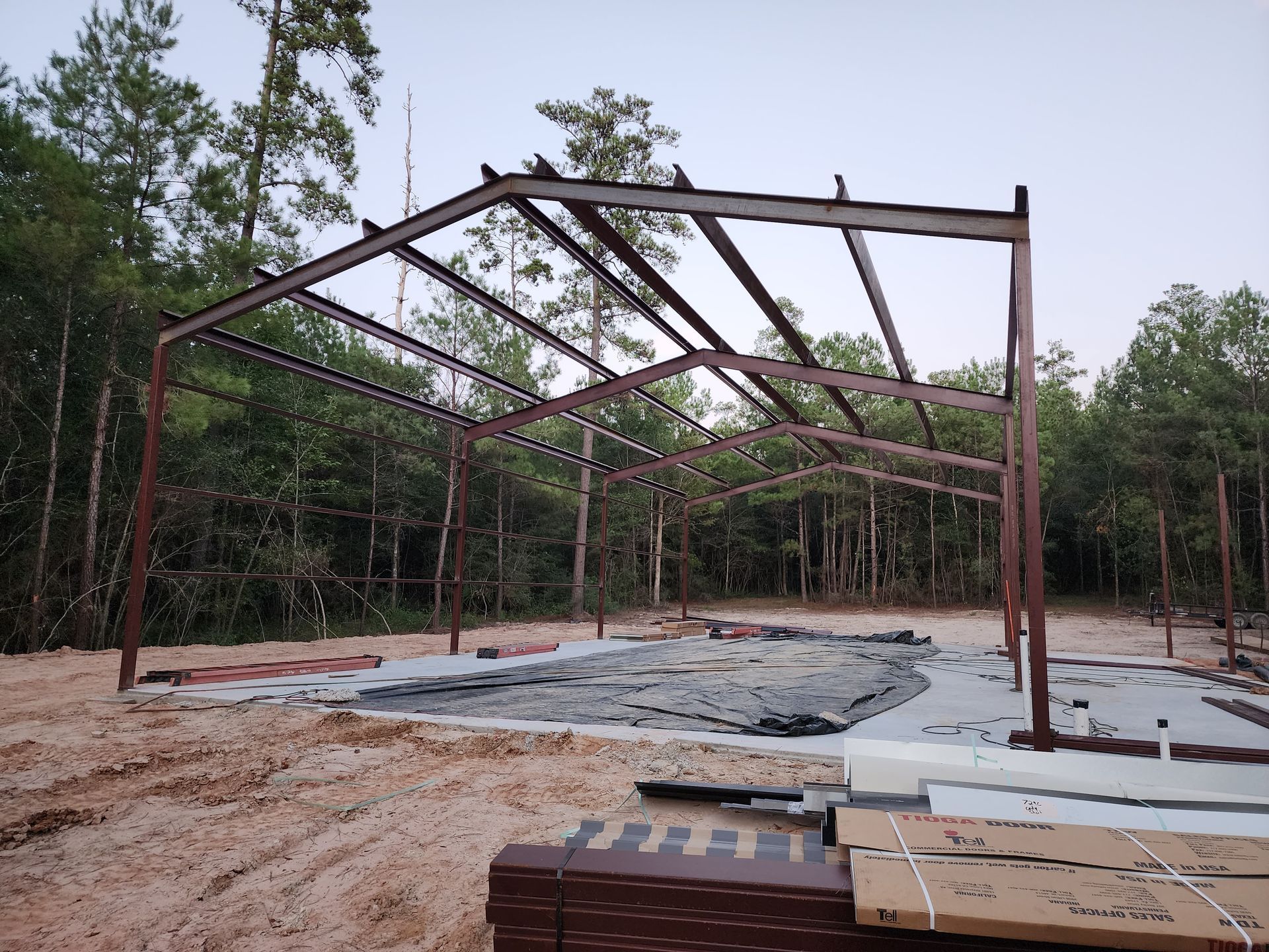 Steel frame of a building under construction in a wooded area, with a concrete foundation and some materials.