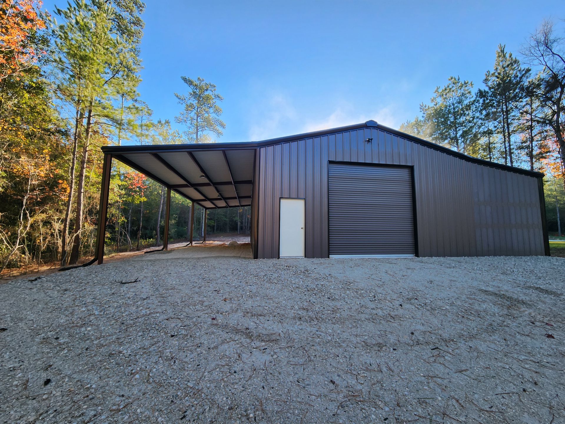 Brown metal barn with carport, gravel ground, and surrounding trees under a blue sky.