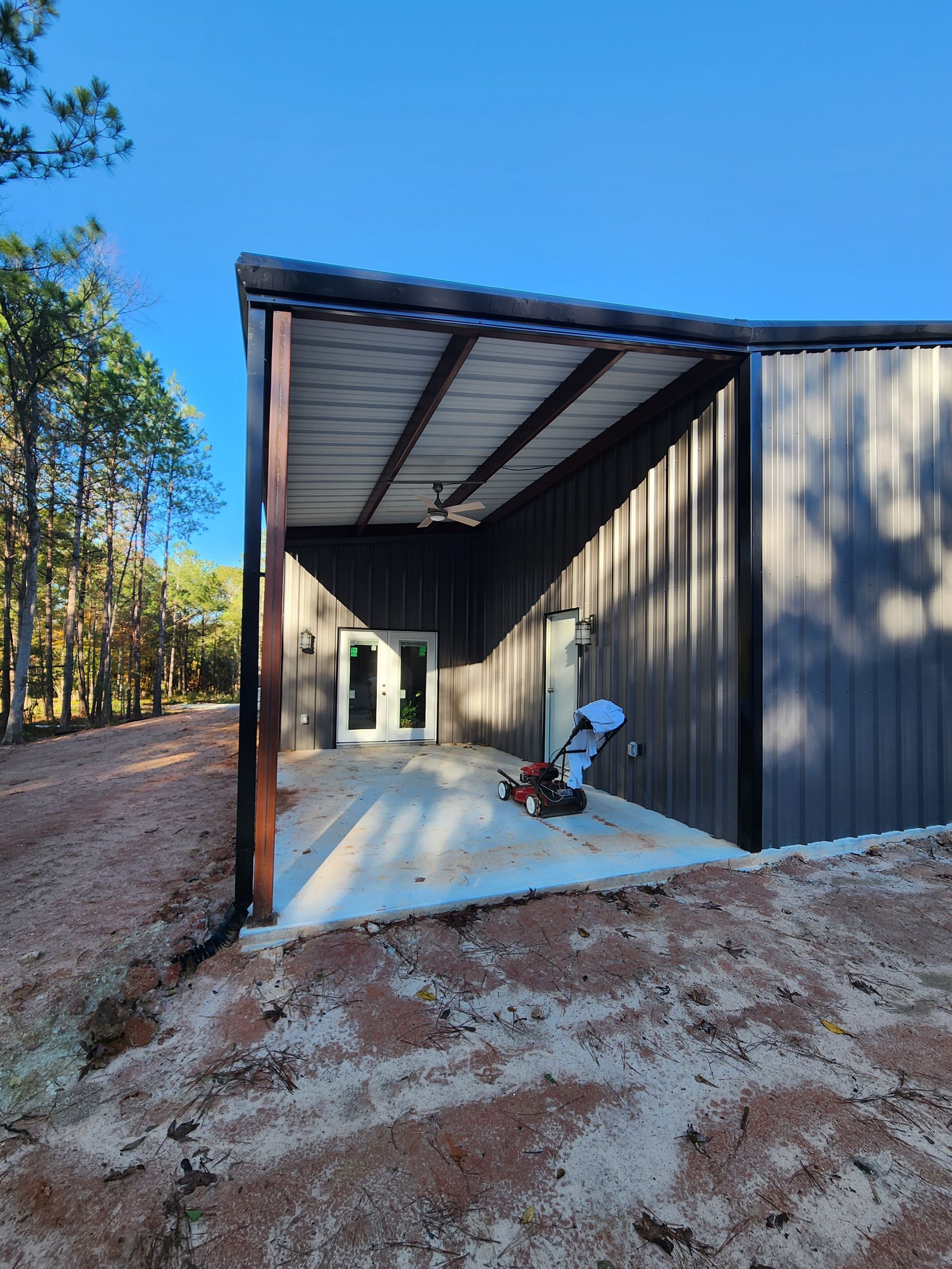 Gray metal building with a concrete patio and brown supports; a lawnmower sits on the patio.