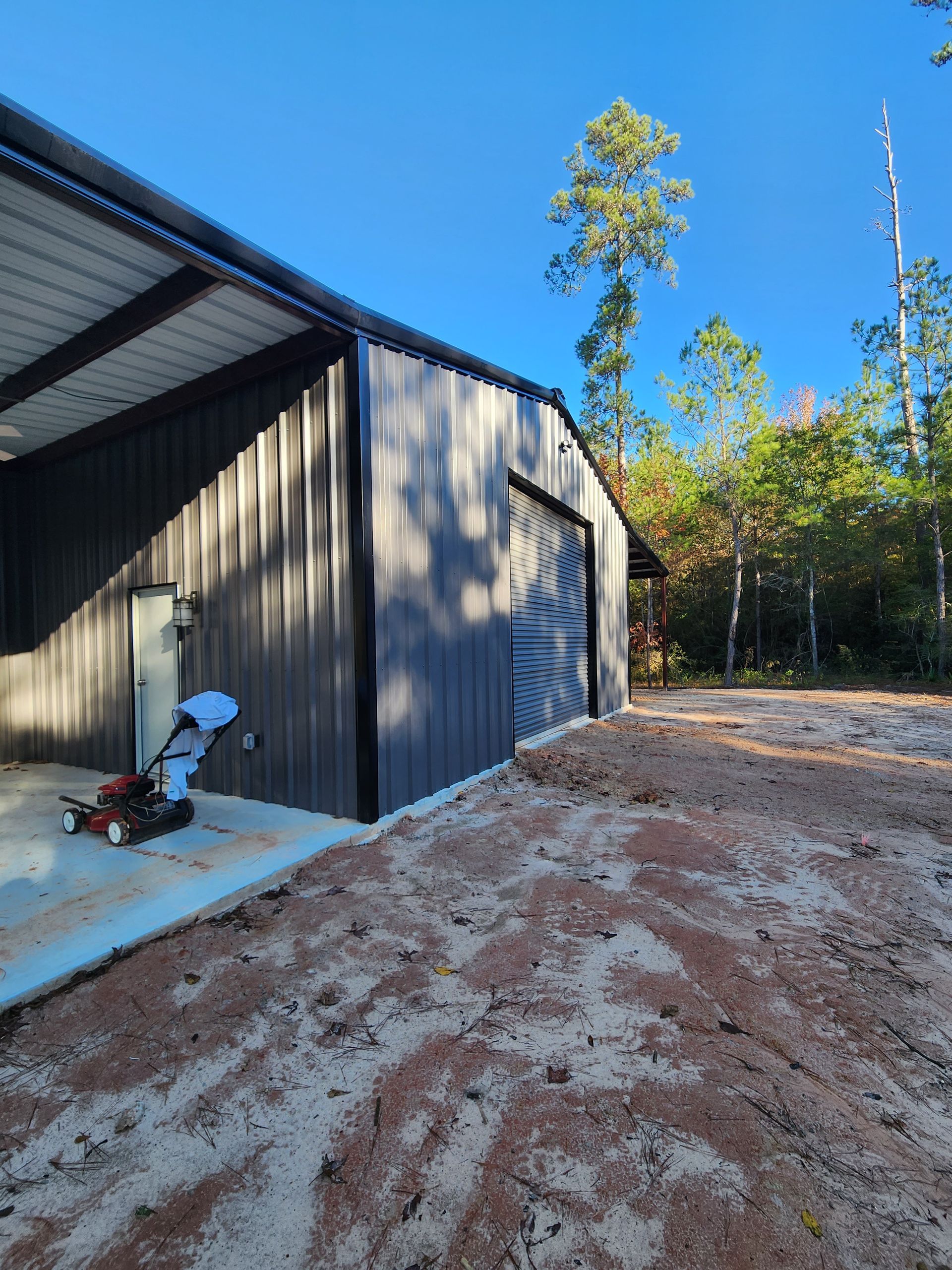 Black metal shed with roll-up door and covered patio, surrounded by dirt and trees, sunny day.