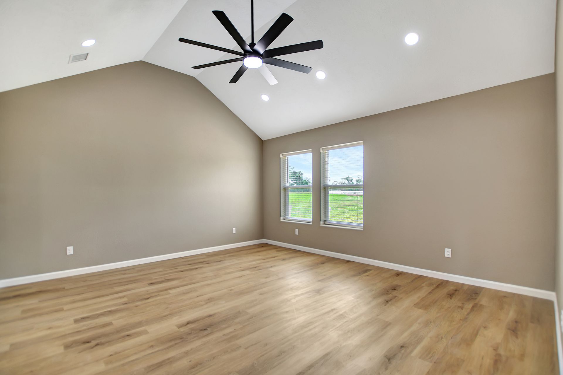 Empty room with vaulted ceiling, light wood floors, and a black ceiling fan. Two windows overlook a green field.