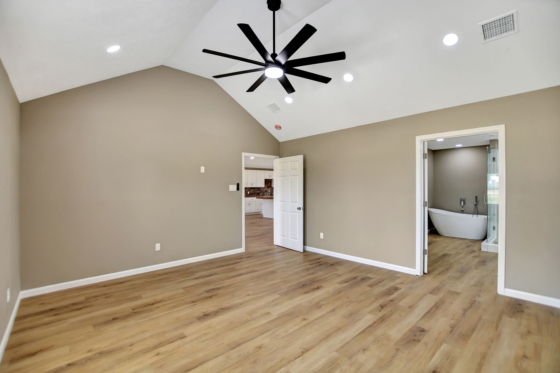 Spacious bedroom with vaulted ceiling, wooden floors, neutral walls, and an open doorway to a bathroom with a tub.