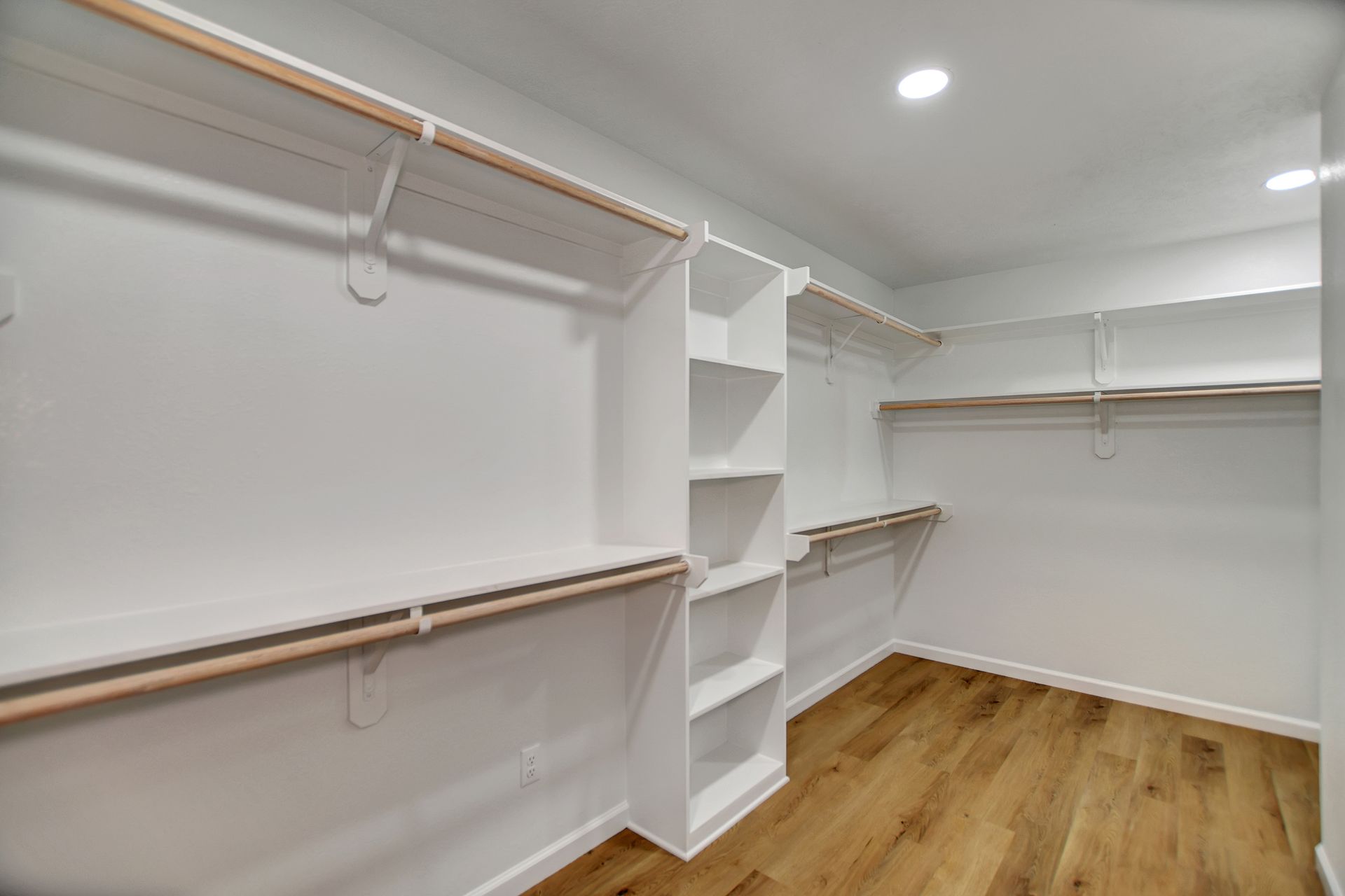 Empty walk-in closet with white shelves and rods, light wood floor, and recessed lighting.