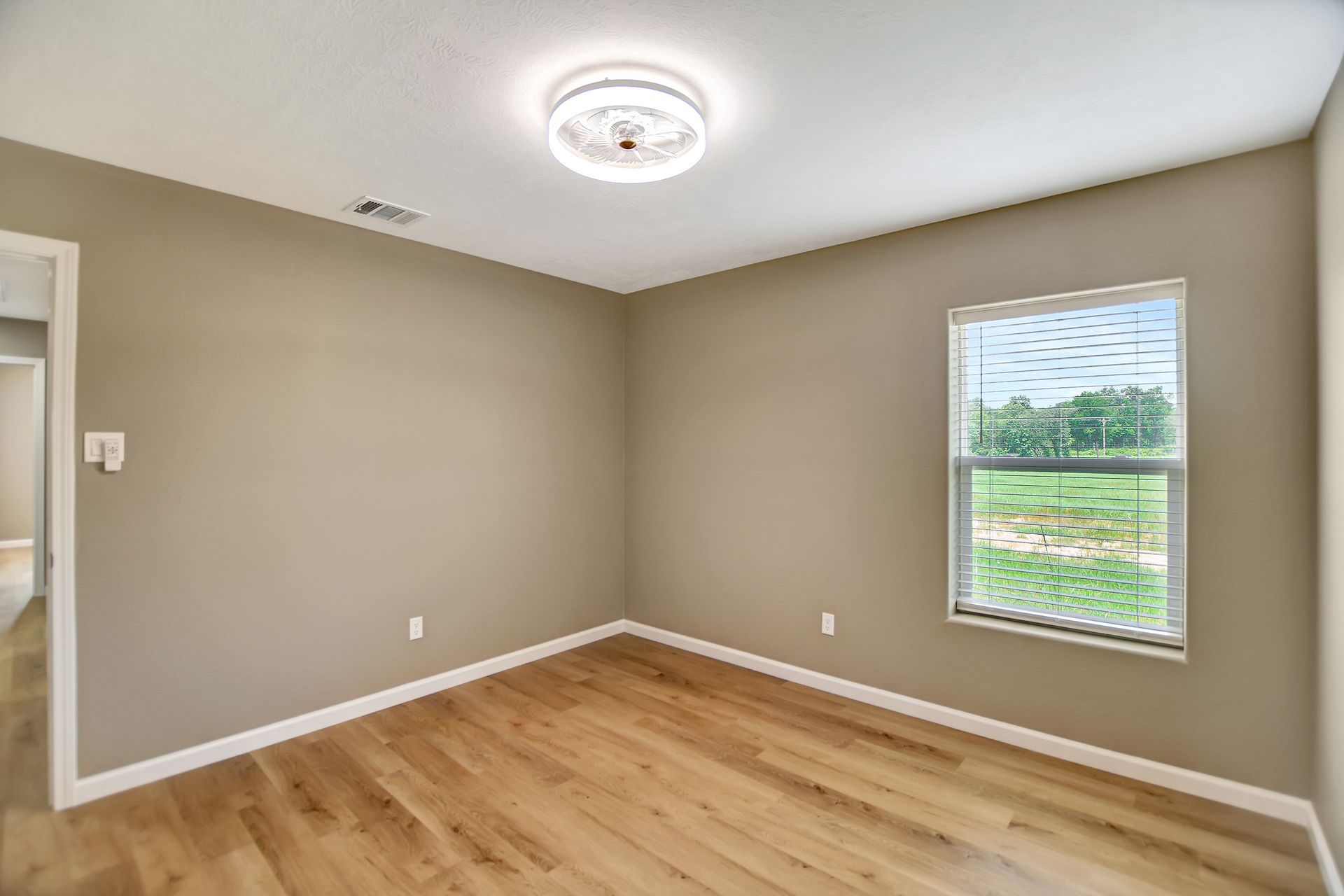 Empty room with beige walls, wooden floor, and a window with a view. Ceiling fan light fixture.