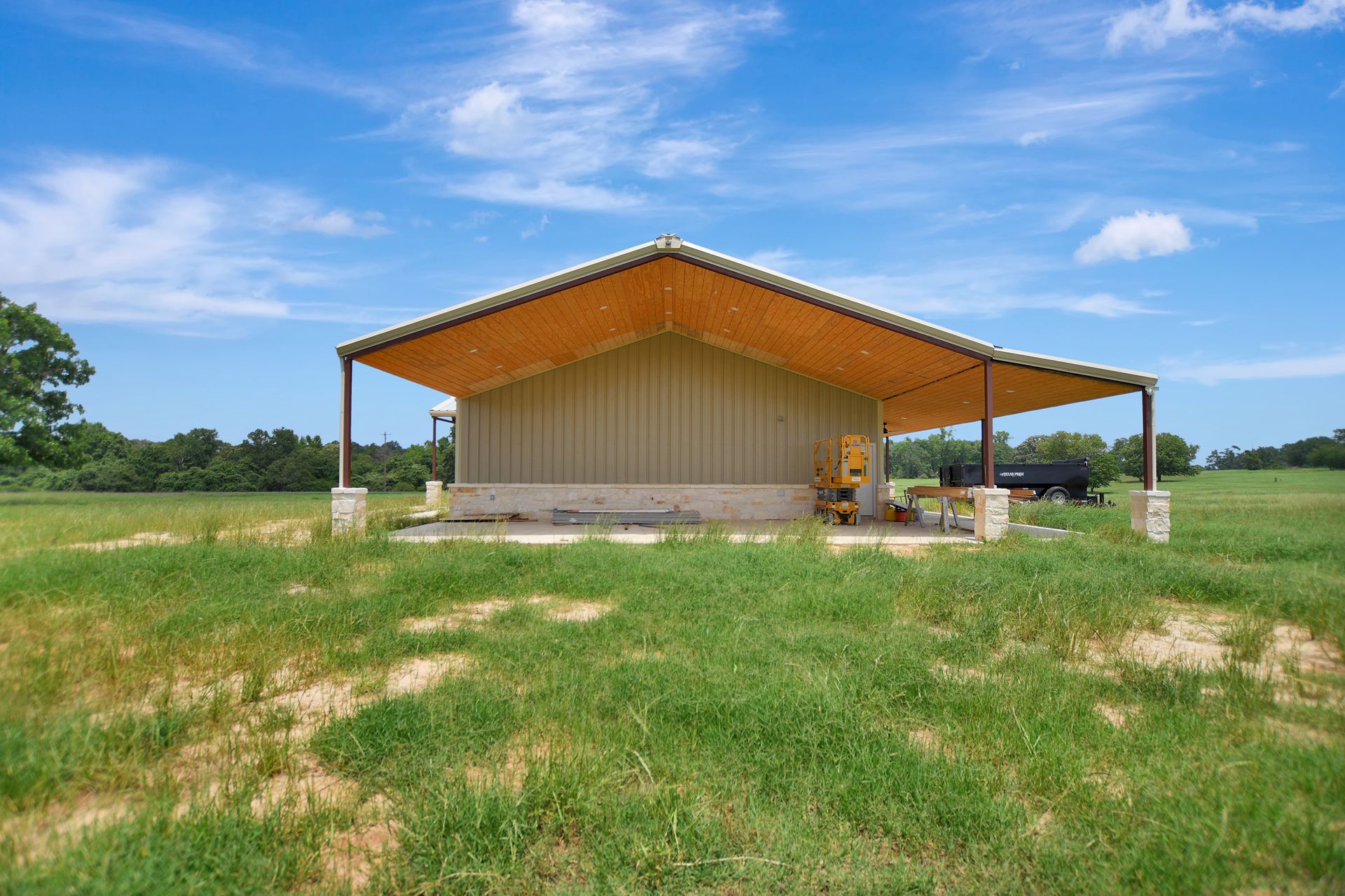 A metal-roofed shelter with concrete columns on a grassy field under a blue sky.