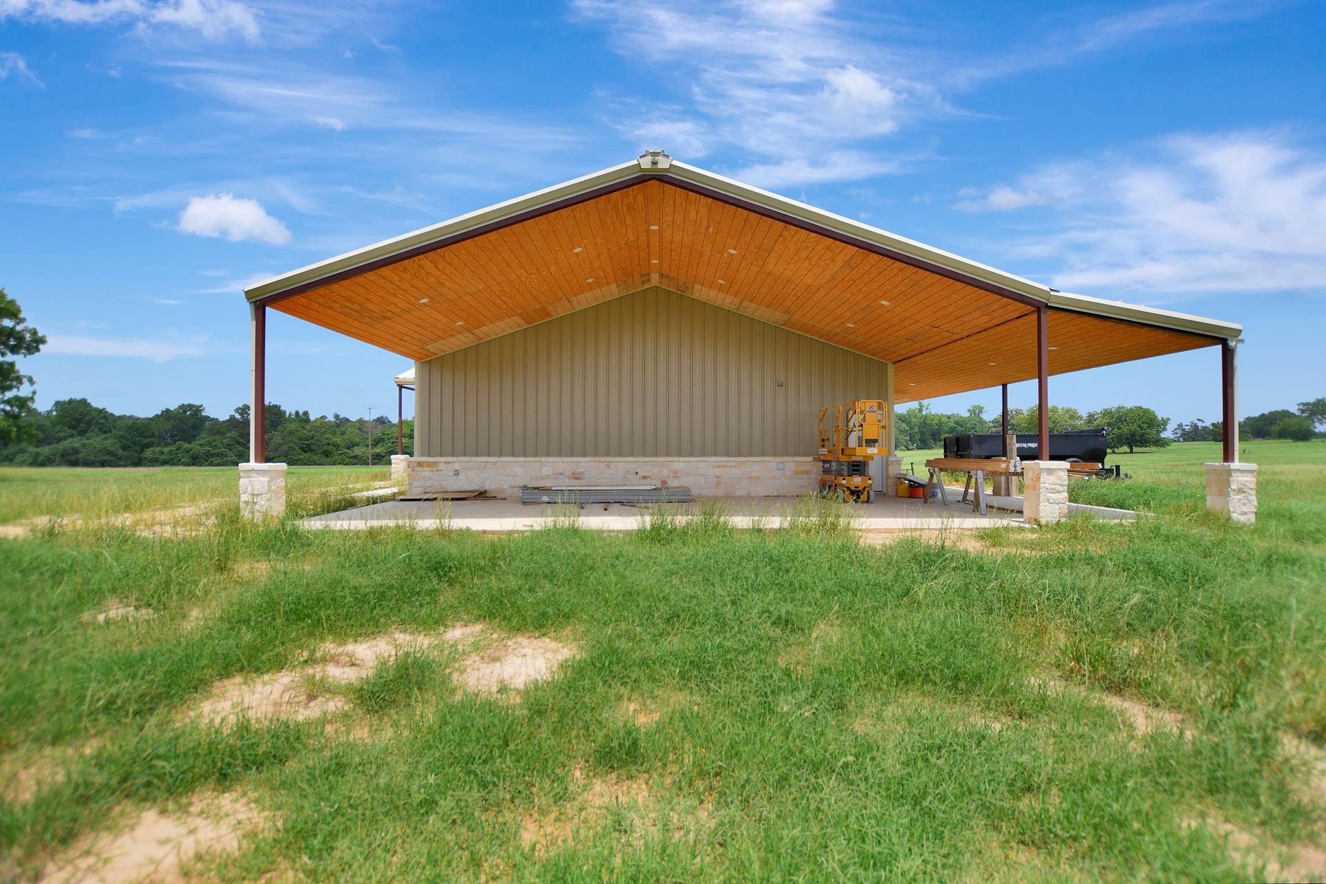 Metal barn with tan roof and siding, brown support beams, set on a concrete pad in a grassy field under a blue sky.
