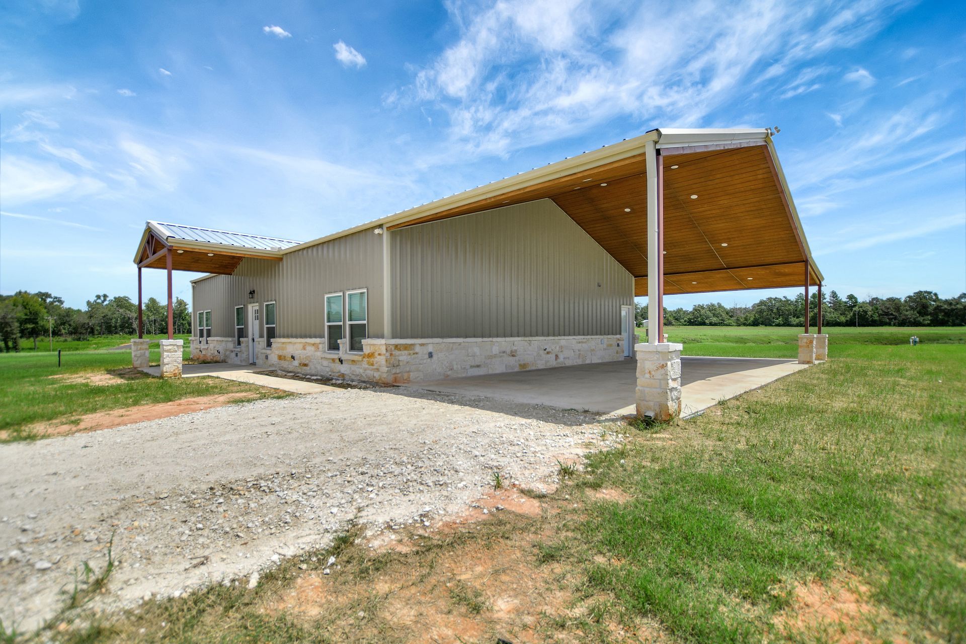 Metal-sided house with a large carport, gravel driveway, and green fields under a blue sky.