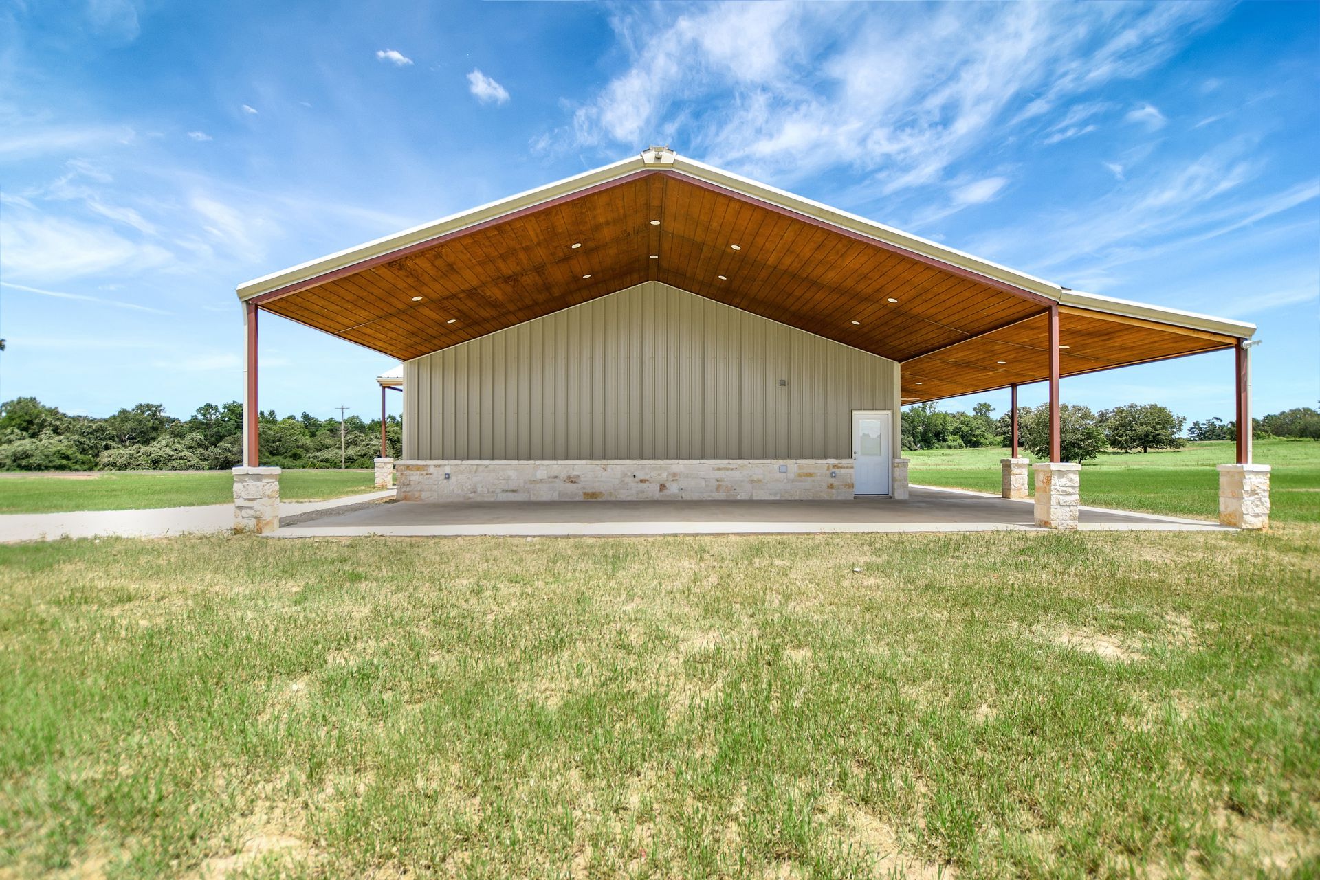 Tan metal building with an extended awning supported by red pillars, set in a grassy field under a blue sky.