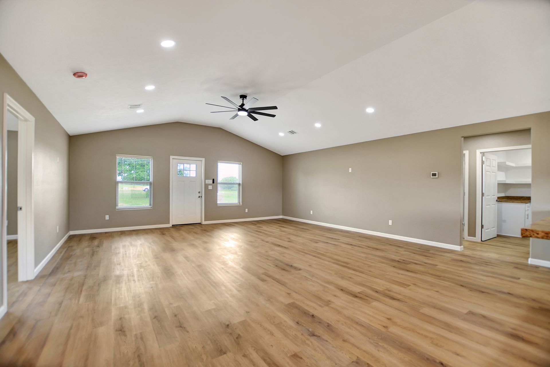 Spacious, empty living room with vaulted ceiling, wood flooring, and neutral walls; door and windows at the back.