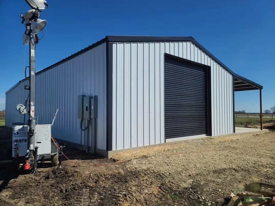 A white metal building with a black garage door is sitting in the middle of a dirt field.