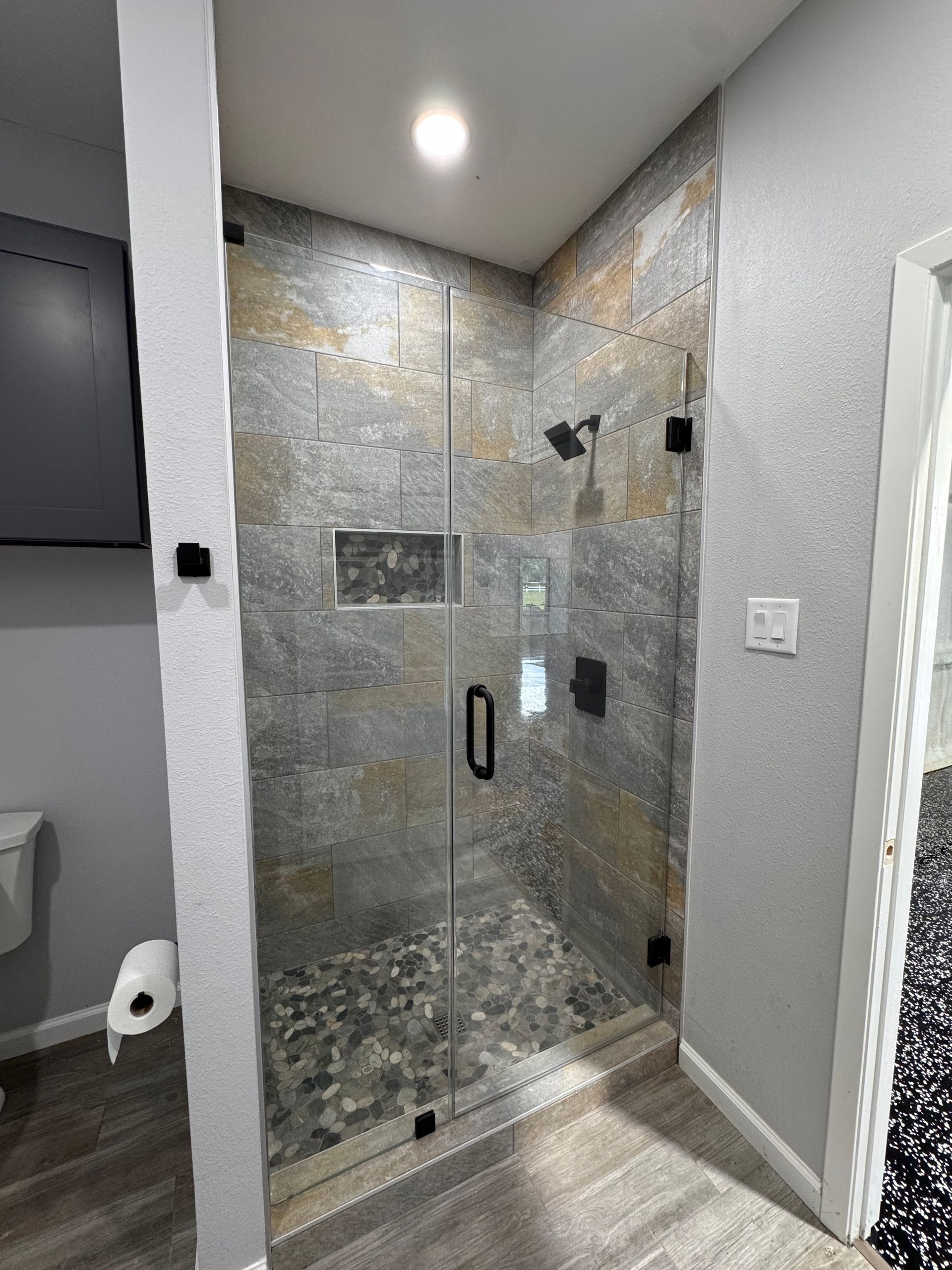 Shower with stone tile walls, glass door, pebble floor, and black fixtures.