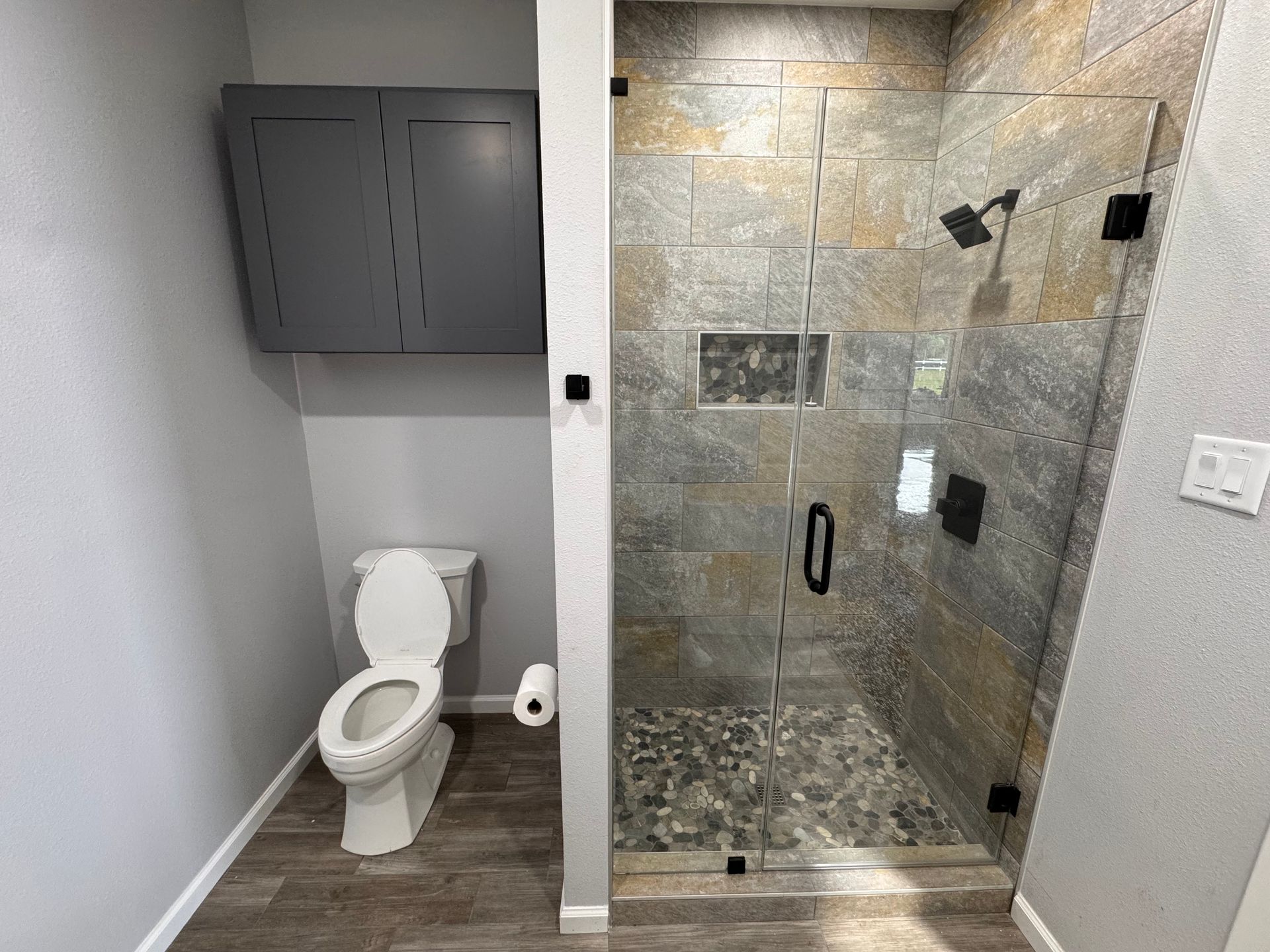 Bathroom with a shower, toilet, and cabinet. Gray and brown tones with glass shower door.