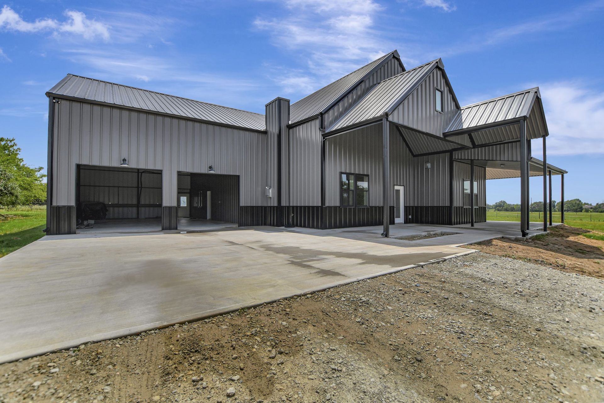 Modern gray metal barn-style house with a concrete driveway and carport under a blue sky.