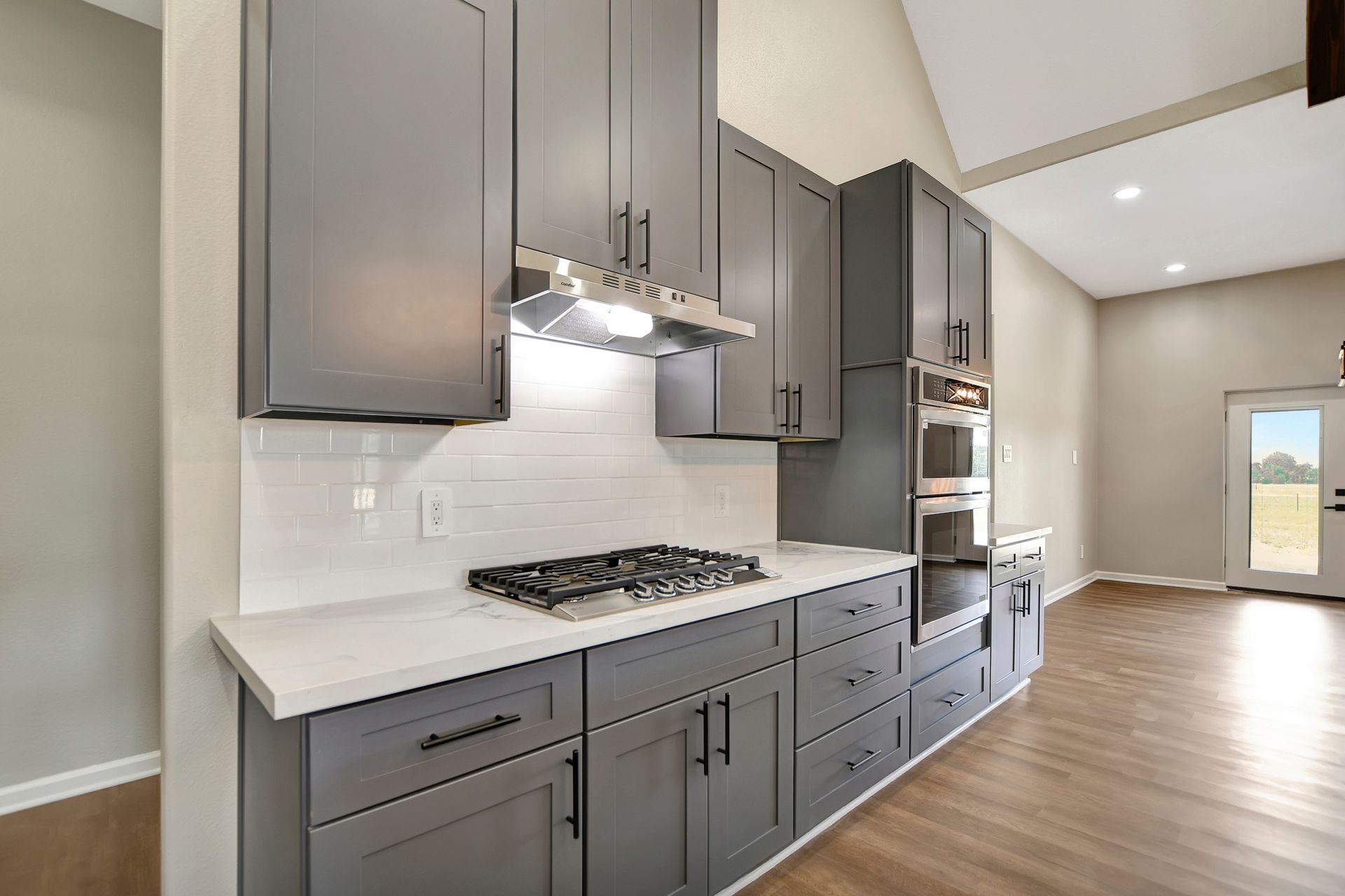 Modern gray kitchen with white countertops, cabinets, and oven, wood floor, and a view of a door.