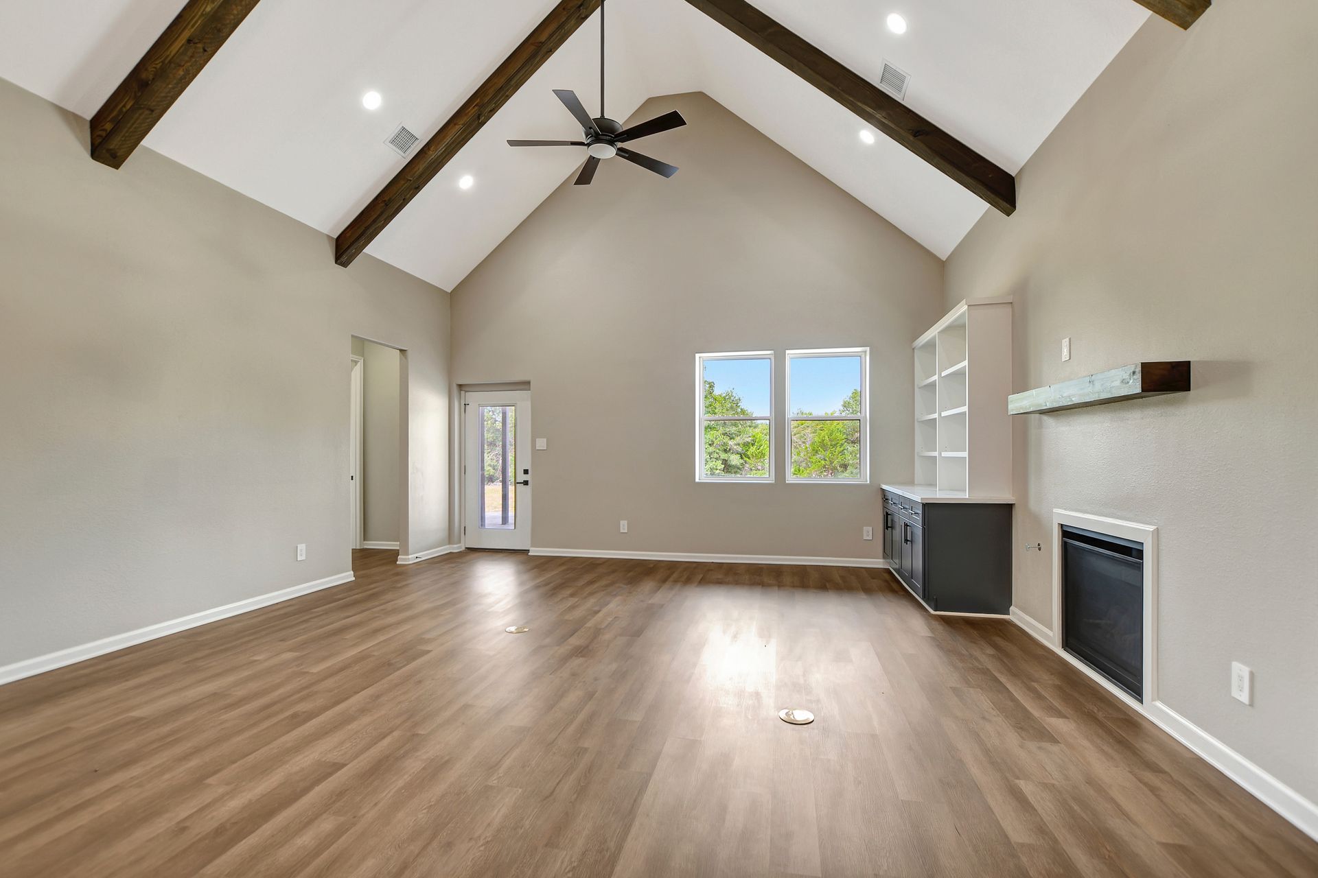Spacious living room with vaulted ceiling, dark wood beams, hardwood floor, and windows.