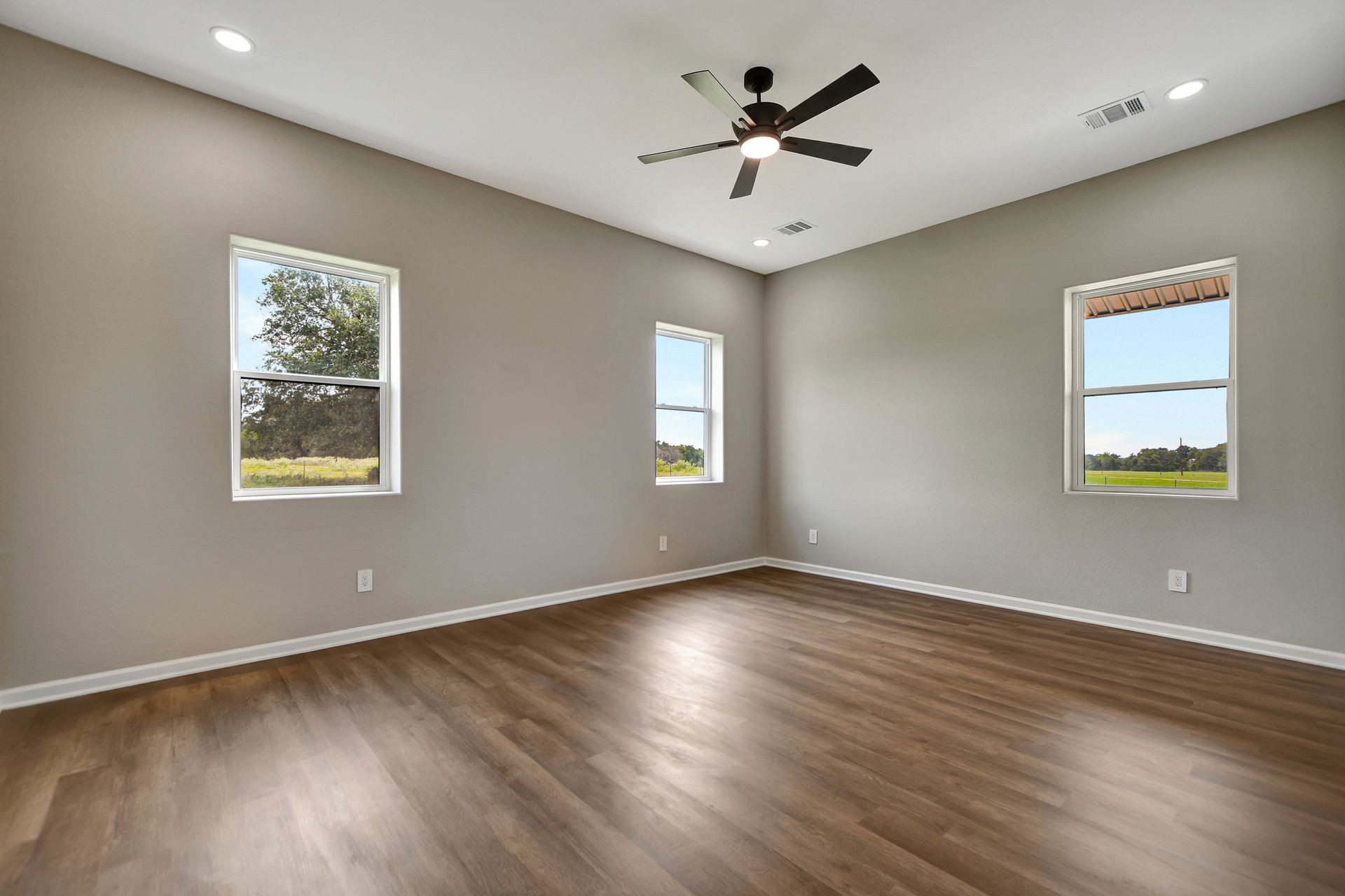 Empty room with three windows, a ceiling fan, and hardwood floors. Gray walls.