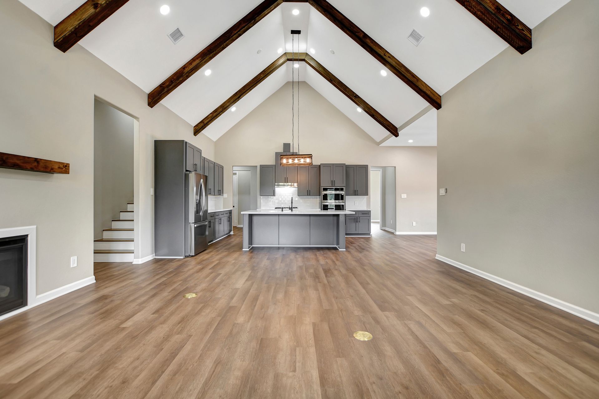 Spacious interior with vaulted ceiling, wood beams, kitchen, and fireplace. Brown floors and gray walls.
