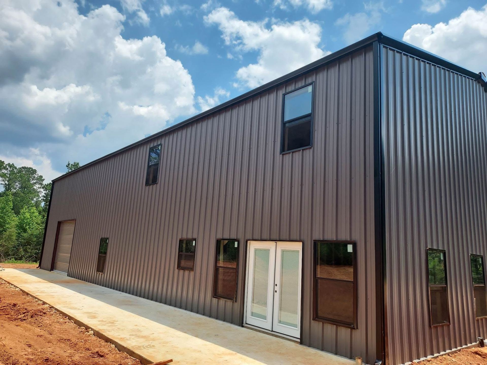 Brown metal building with windows and double doors, concrete walkway, against a cloudy sky.