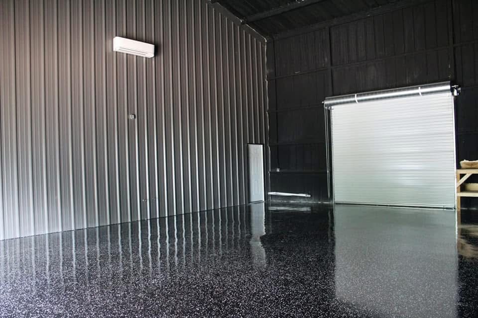 Shiny black epoxy floor inside a metal building with a roll-up door and corrugated silver walls.