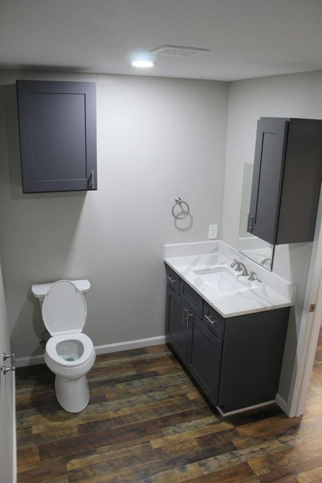 Bathroom with gray cabinets, white toilet and sink, brown flooring, and a towel ring.