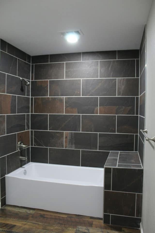 Bathroom with white tub, dark tile walls, and brown flooring.