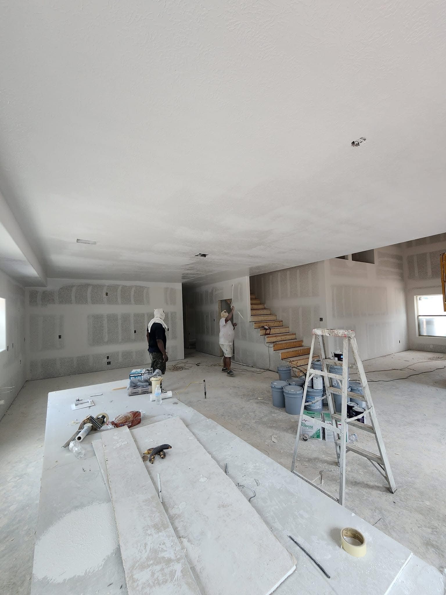 Interior of a house under construction, drywall and unfinished staircase visible. People are present, tools and materials scattered.
