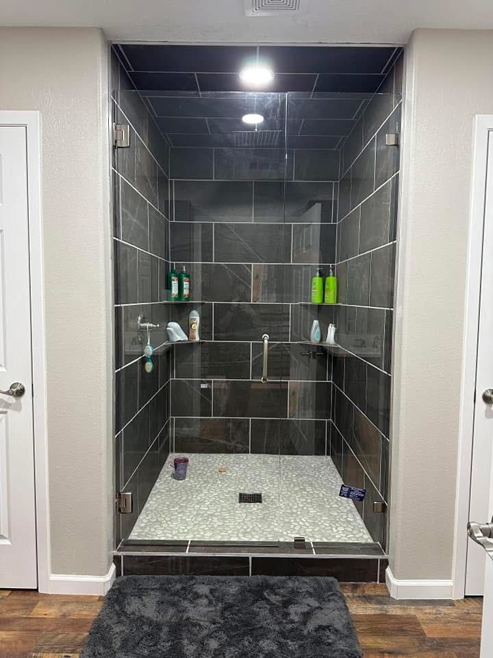 Modern walk-in shower with dark tiled walls, glass doors, and stone floor, set between two white doors, with a dark gray rug.