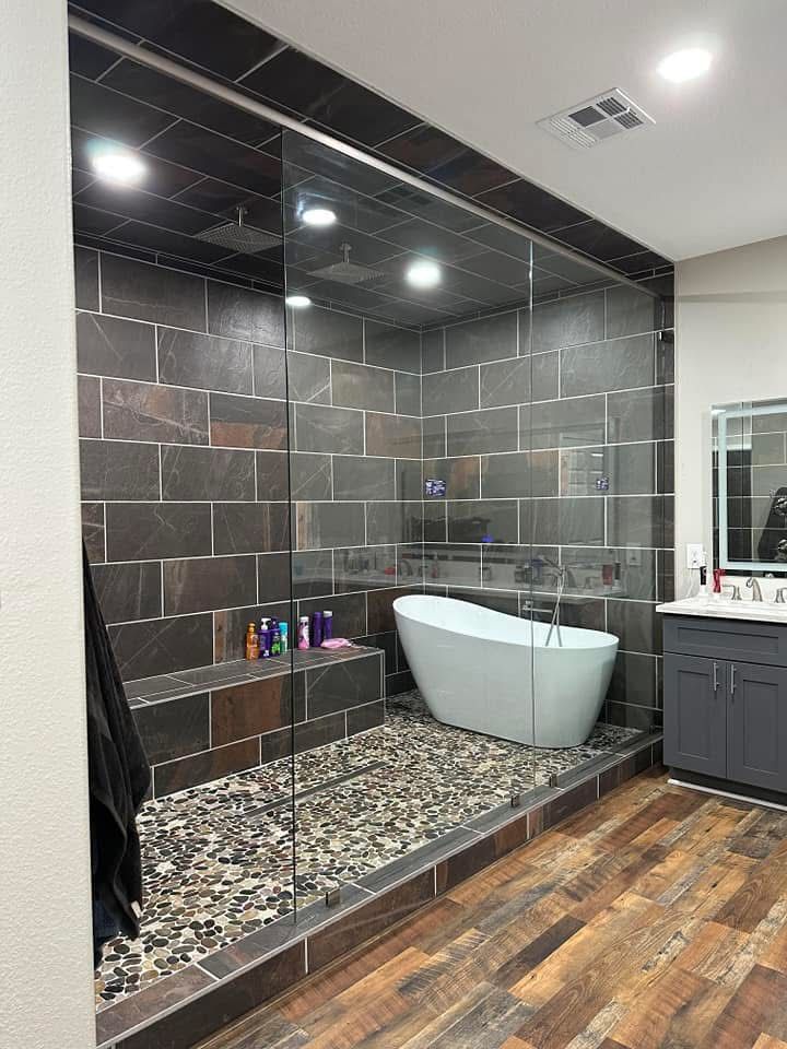 Spacious modern bathroom with a glass shower and a freestanding tub; dark tile walls, stone floor, and wood flooring.