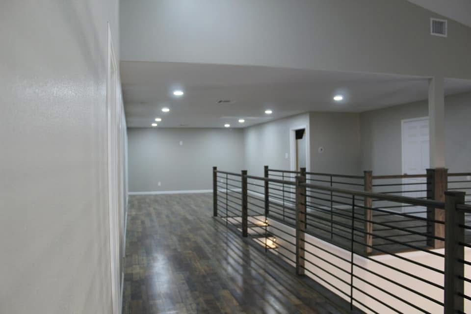 A long, empty room with gray walls and dark flooring, featuring a black railing and recessed ceiling lights.
