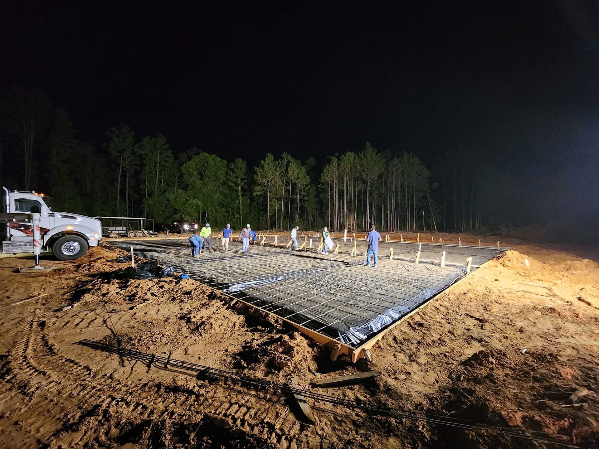 Construction workers pouring concrete at night. Ground level, trees in background. Truck on left.