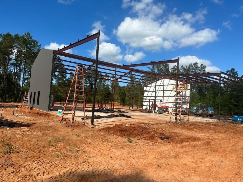 Steel frame of a building under construction with a partially completed wall against a blue sky and trees.