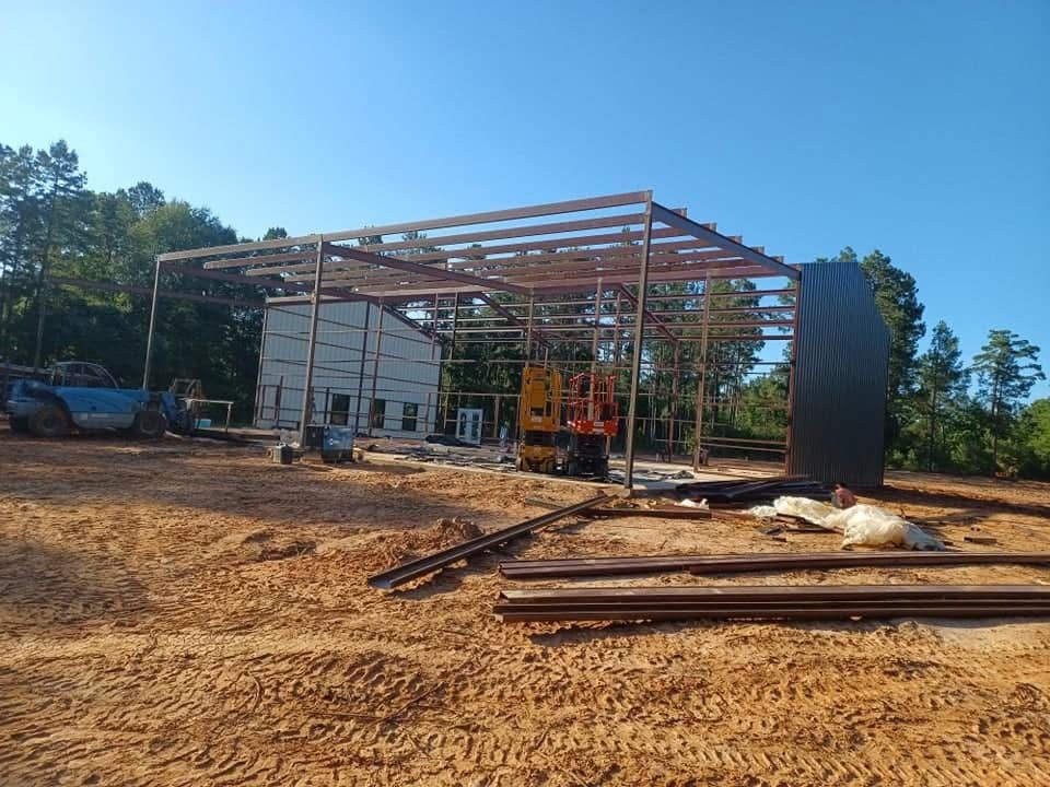 Steel building under construction on a dirt lot; dark metal siding sits beside the frame, sunny day.