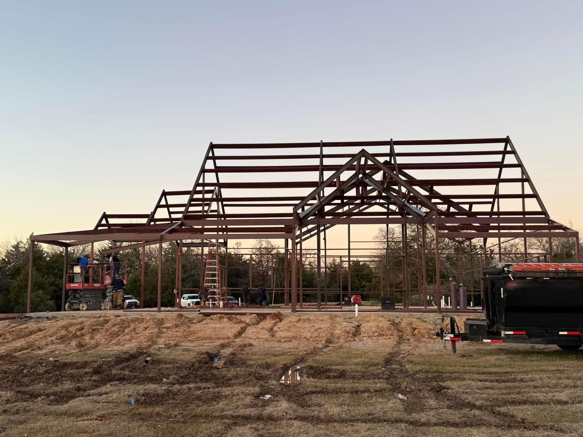 Steel framework of a building under construction. Workers on lifts, trees in background, brown earth.