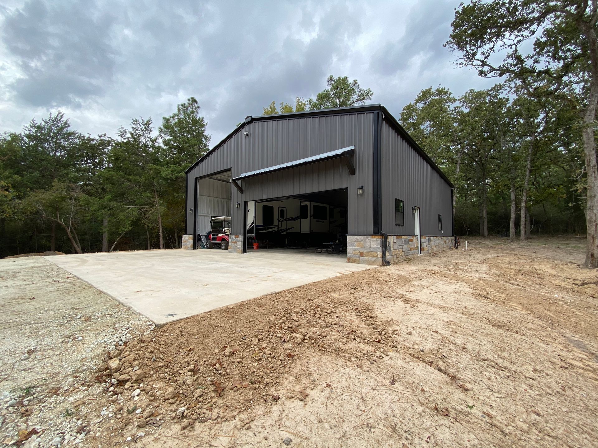 Gray metal building with a concrete pad in a wooded area. The open door reveals a recreational vehicle.