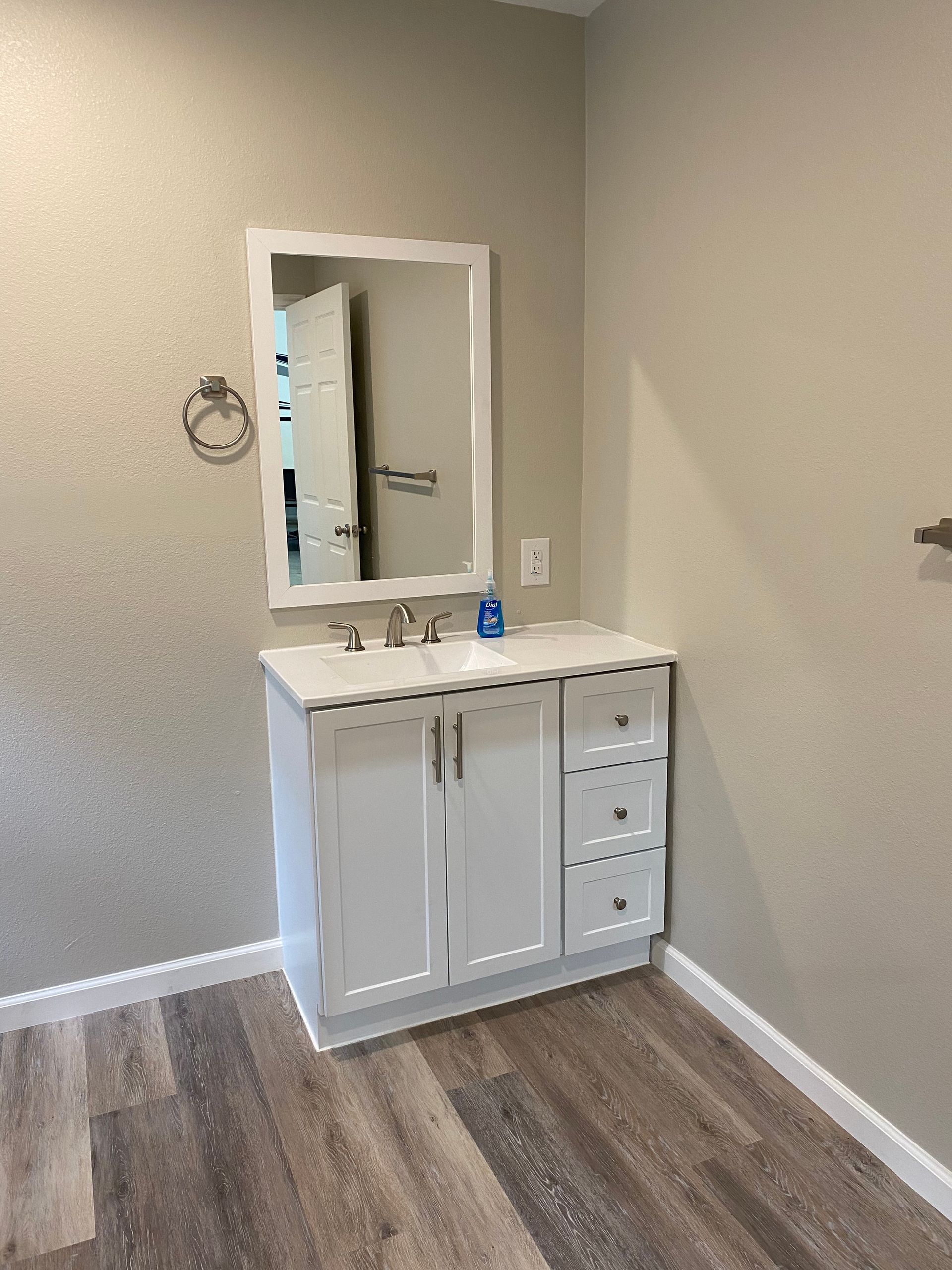 White bathroom vanity with a mirror, towel ring, and drawers, set against beige walls and wood-look flooring.