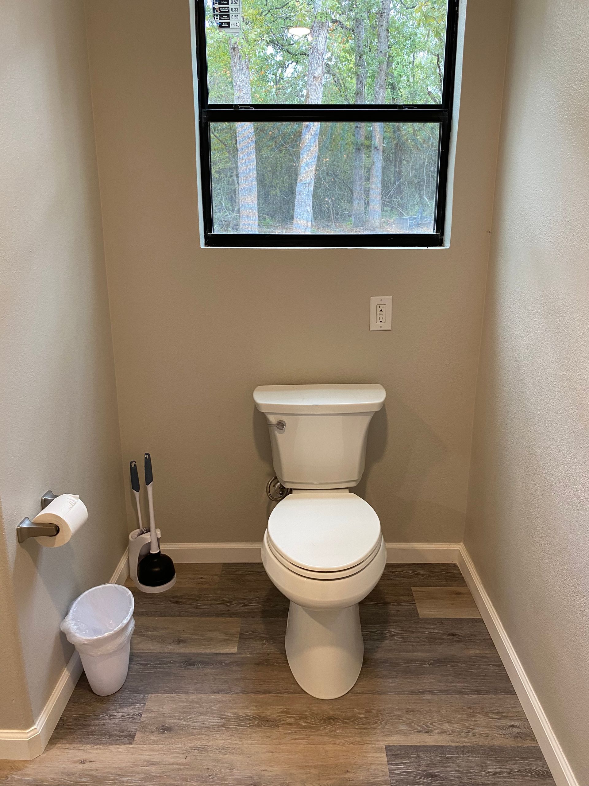 A small bathroom with a toilet, window, and accessories; beige walls, wood-look floor.