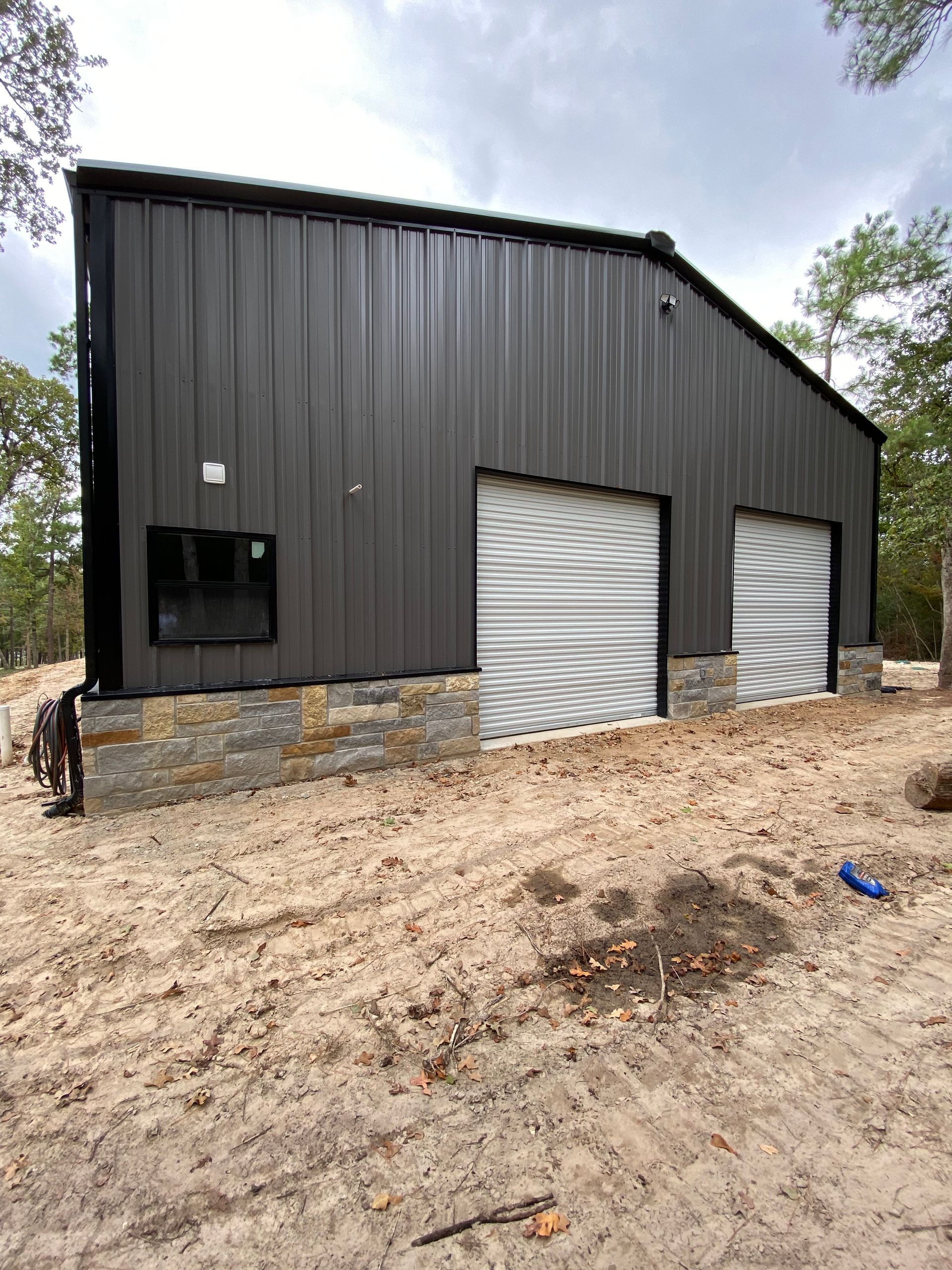 Dark metal building with two garage doors, a small window, and stone base. Located outdoors.