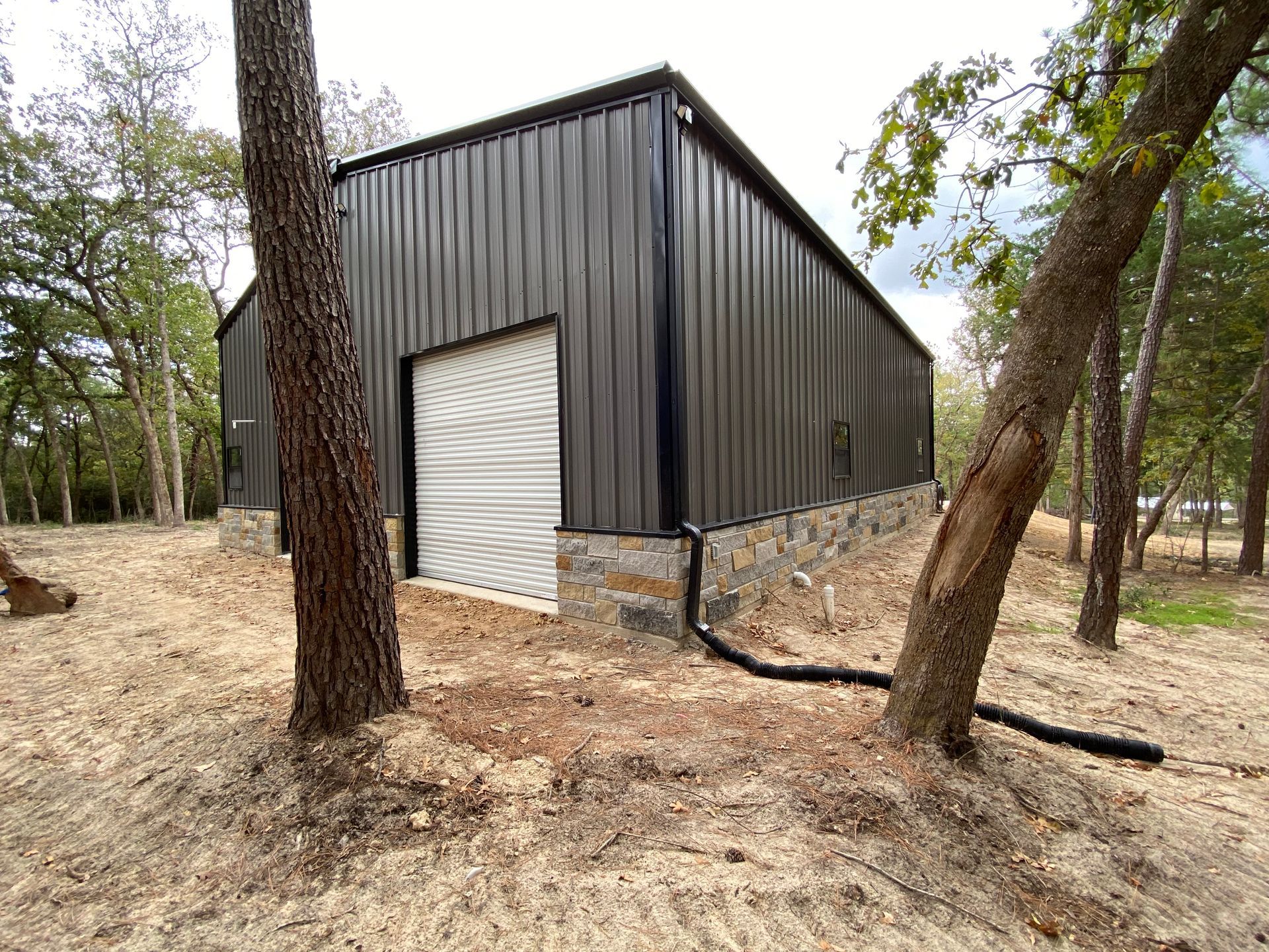 Dark gray metal barn with a stone base in a wooded area.
