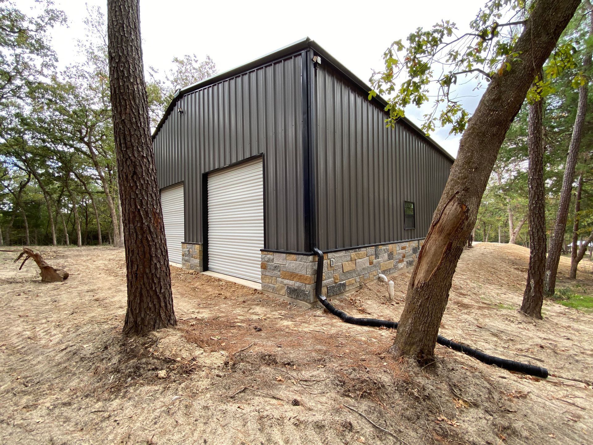 A dark gray metal barn with stone base, surrounded by trees on a sandy ground.