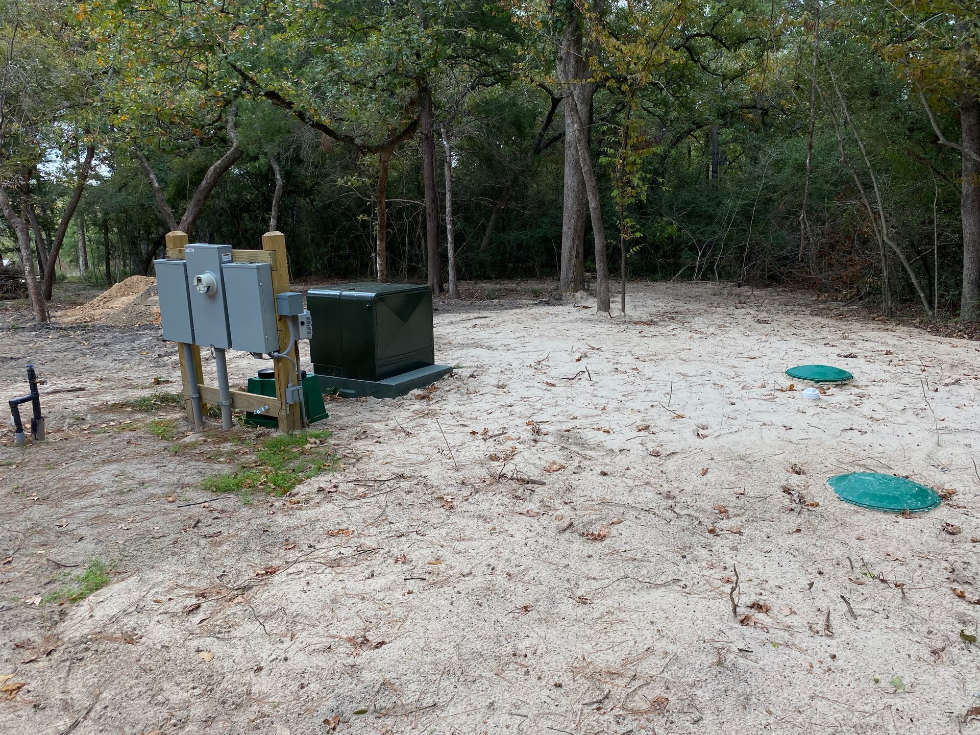 A sand-covered area with green septic tank covers, electrical boxes, and surrounded by trees.