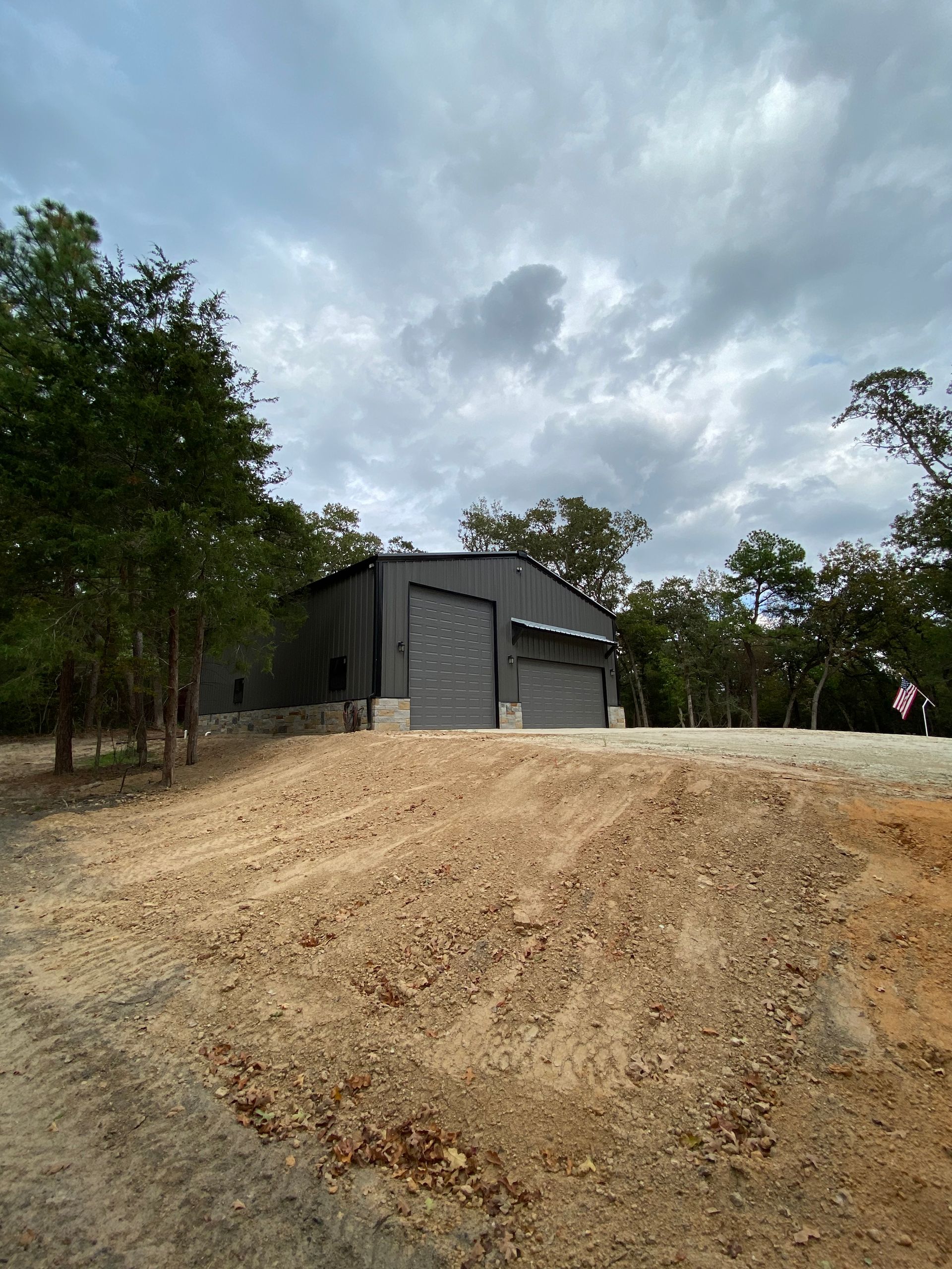 Gray metal building with two garage doors on a gravel hill under cloudy sky.