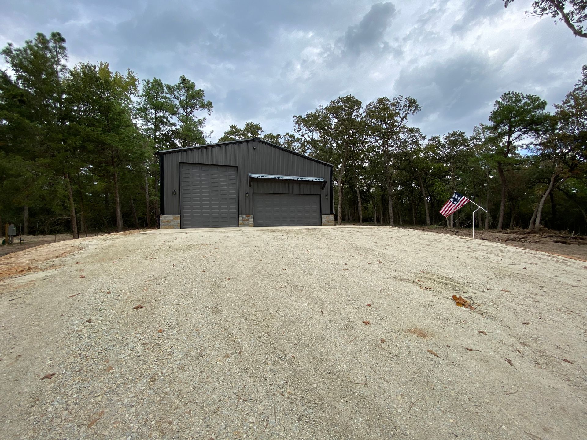 Gray metal building with two garage doors, gravel driveway, trees in the background, cloudy sky, American flag.