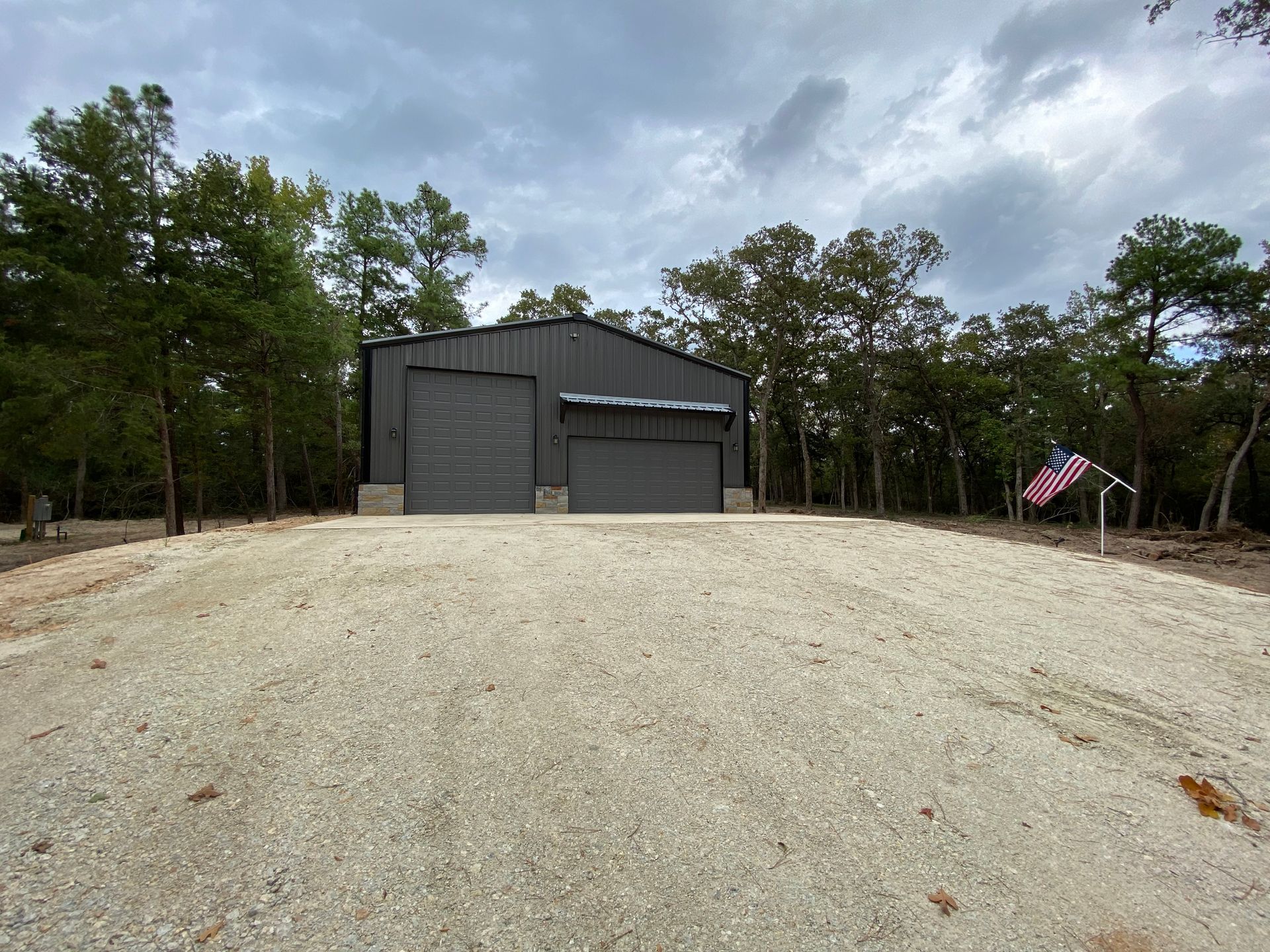 Gray metal building with two garage doors on a gravel drive, American flag waving.