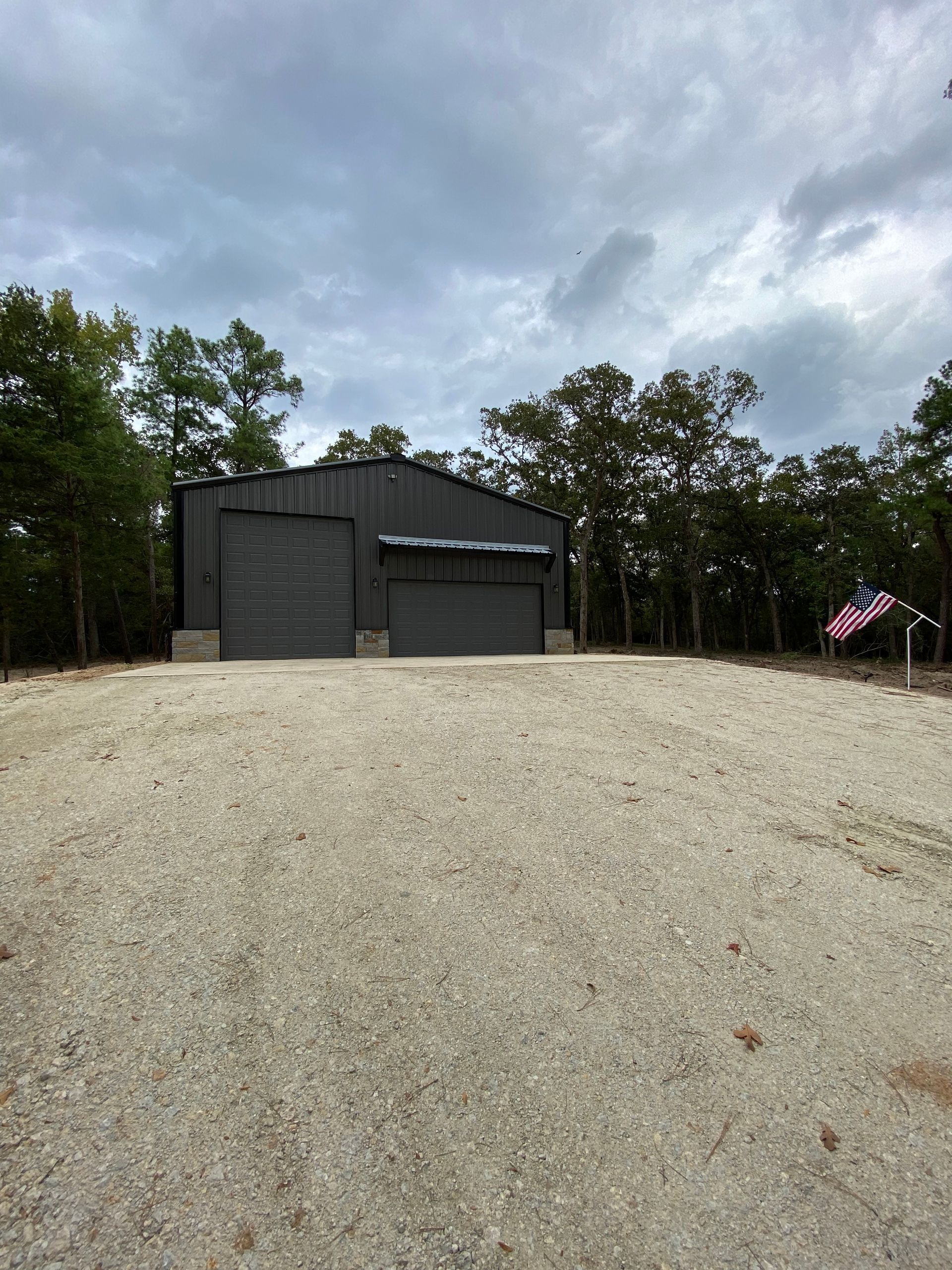 Dark gray metal garage with two bays on gravel lot; surrounded by trees, cloudy sky.