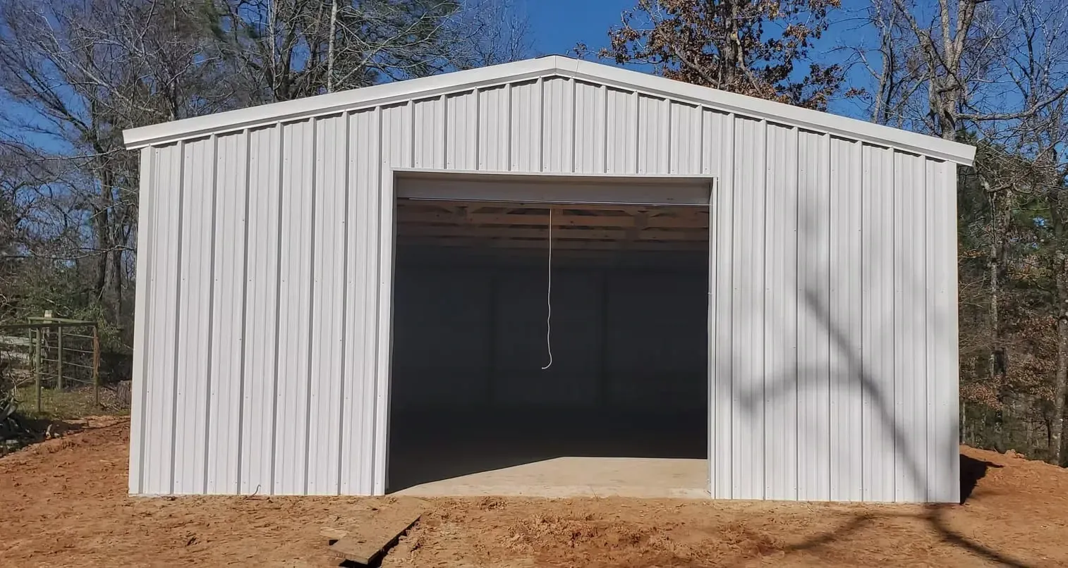 A white metal building with a large door is sitting in the middle of a dirt field.