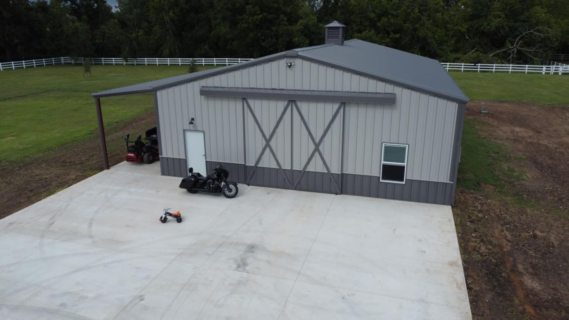 A gray metal barn with a concrete pad. A motorcycle, riding lawnmower, and a small vehicle are in the foreground.