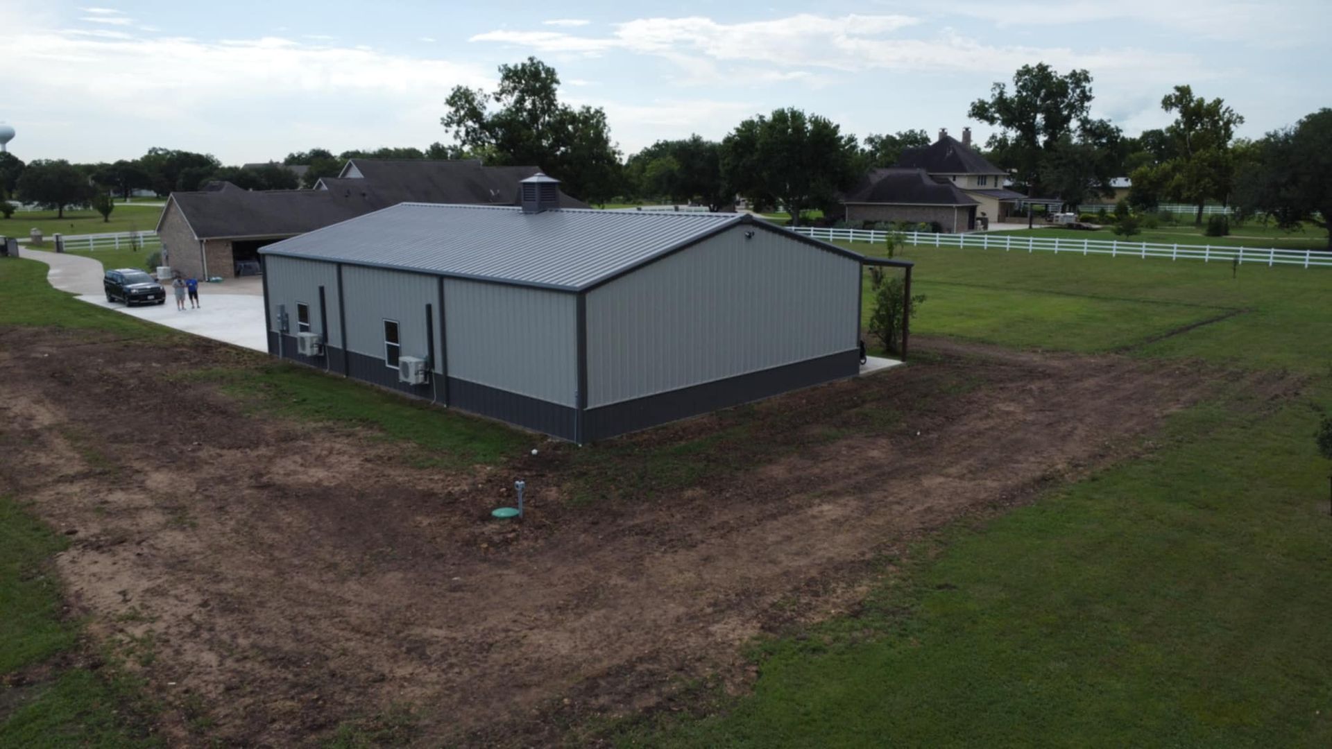 Grey metal barn with dark base, next to a house on grassy property.