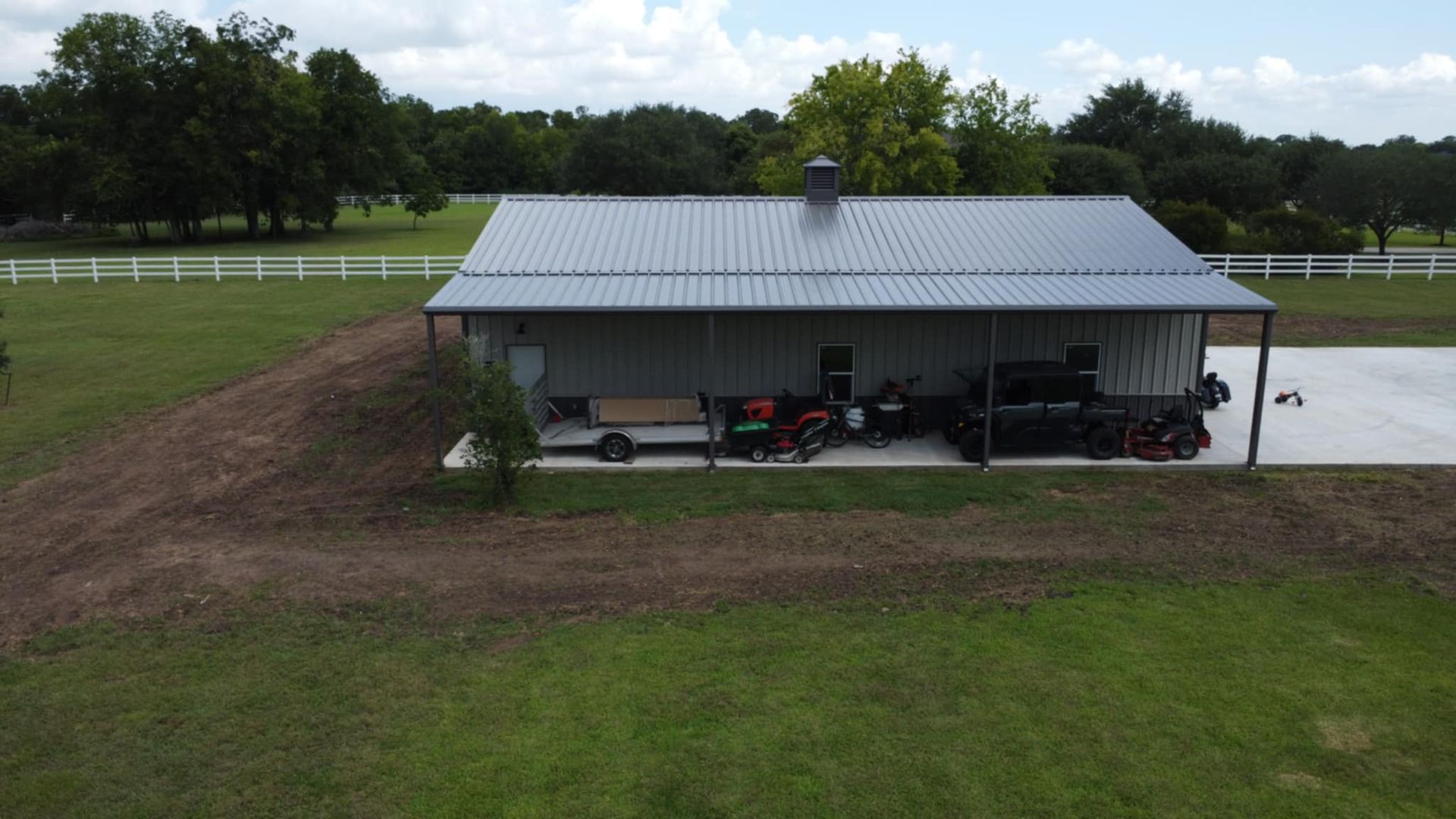 Metal-roofed garage with a concrete apron, motorcycles parked beneath a covered area, and a grass field setting.